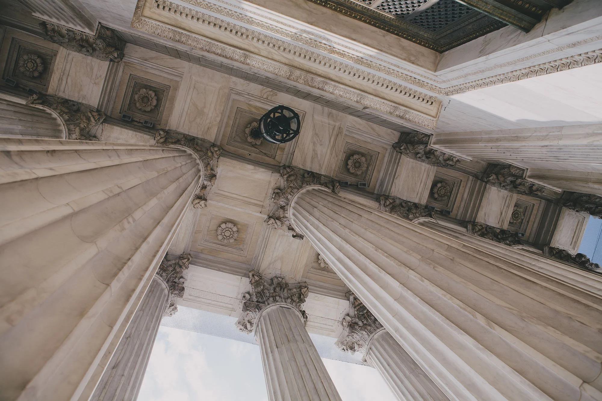 Looking up at marble columns and ceiling with ornate decor at the top of a classical building, Sunlight streams in from above.