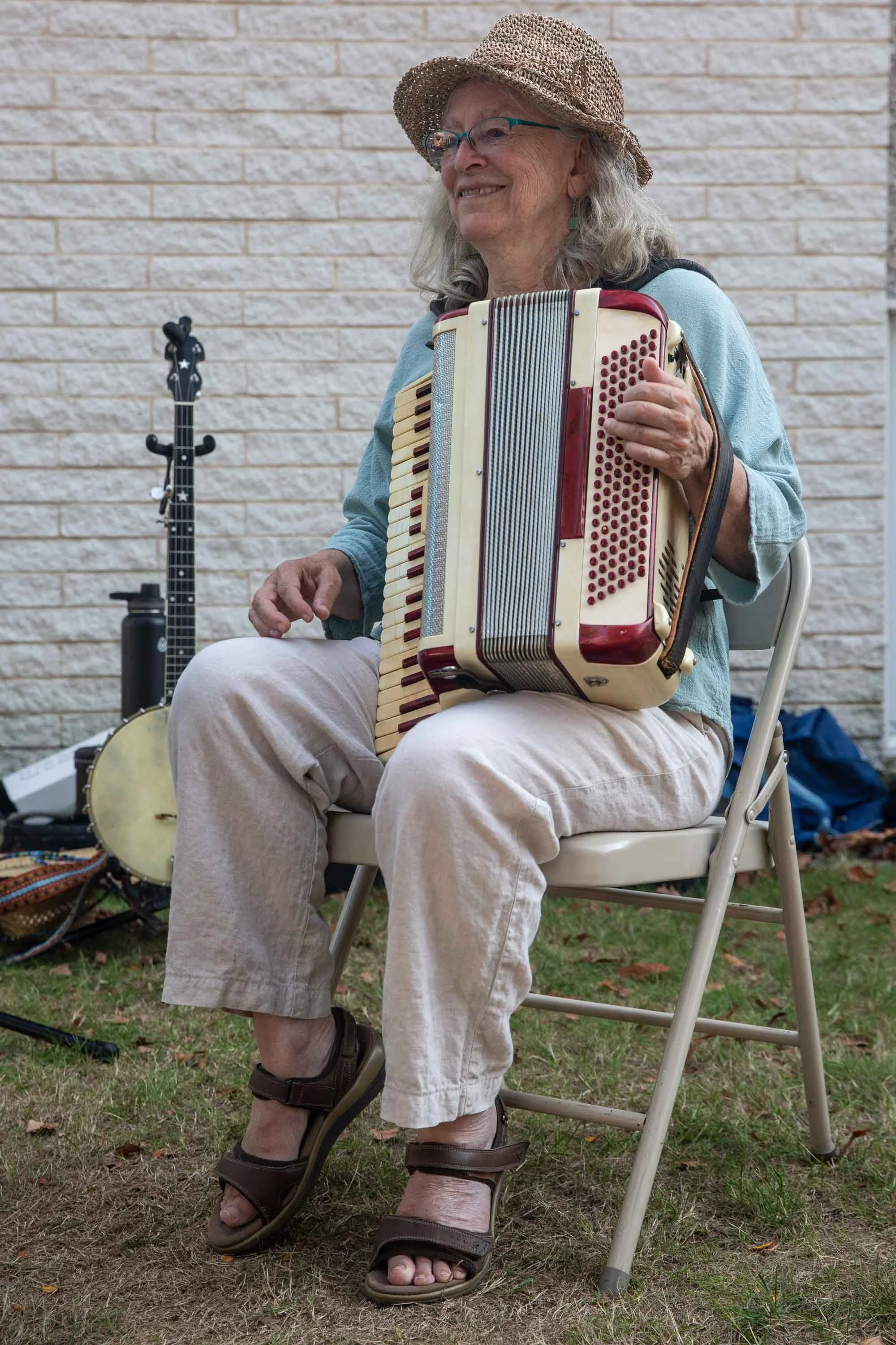 An elderly woman with gray hair, glasses, and a straw hat is sitting on a white folding chair outdoors. She is playing an accordion and smiling. A banjo and an acoustic guitar are visible in the background, along with a brick wall and grass ground.
