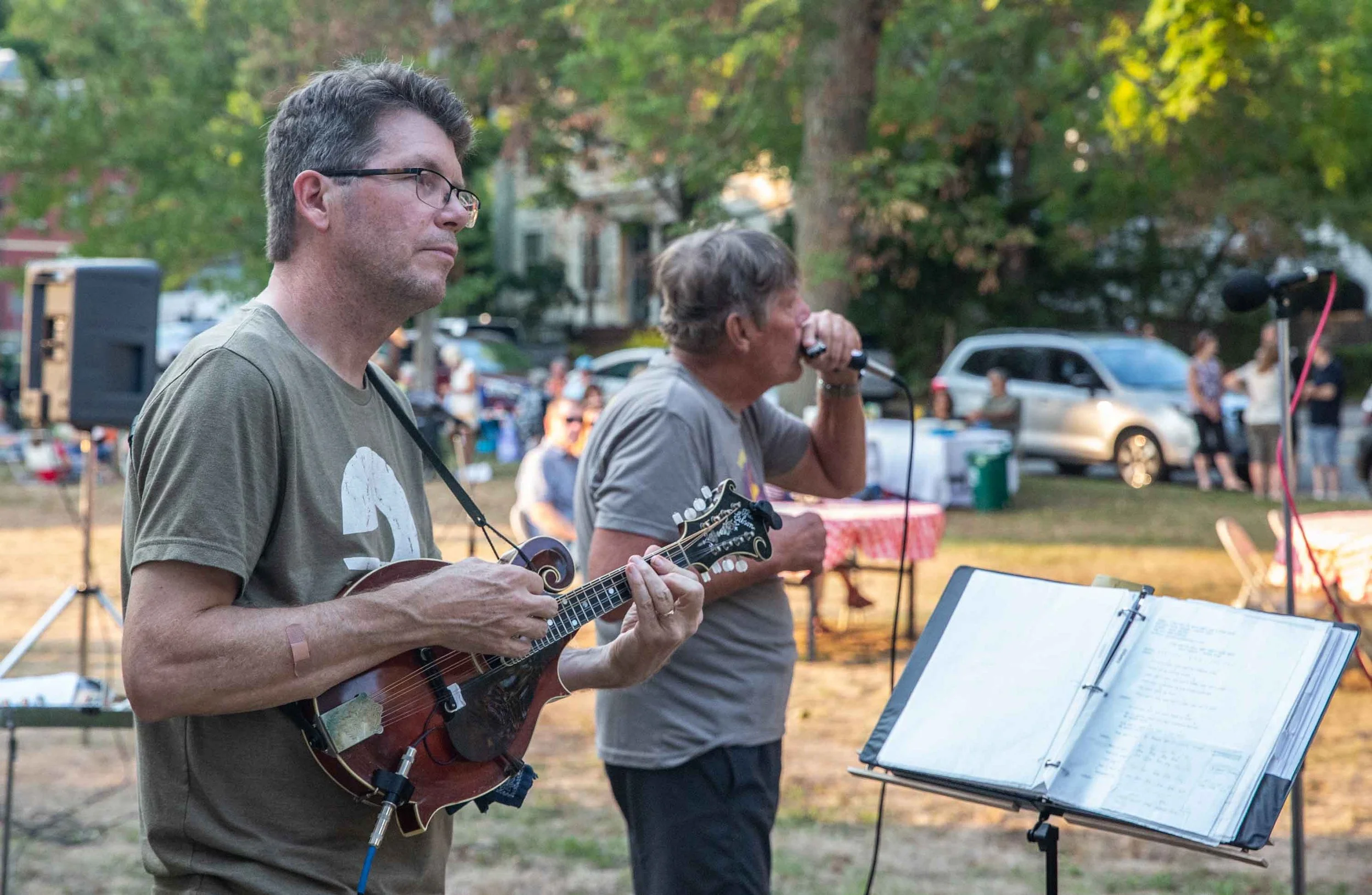 Two men are playing guitars and singing at an outdoor event during the evening. There are music stands with sheet music, and the background shows trees, cars, and people.
