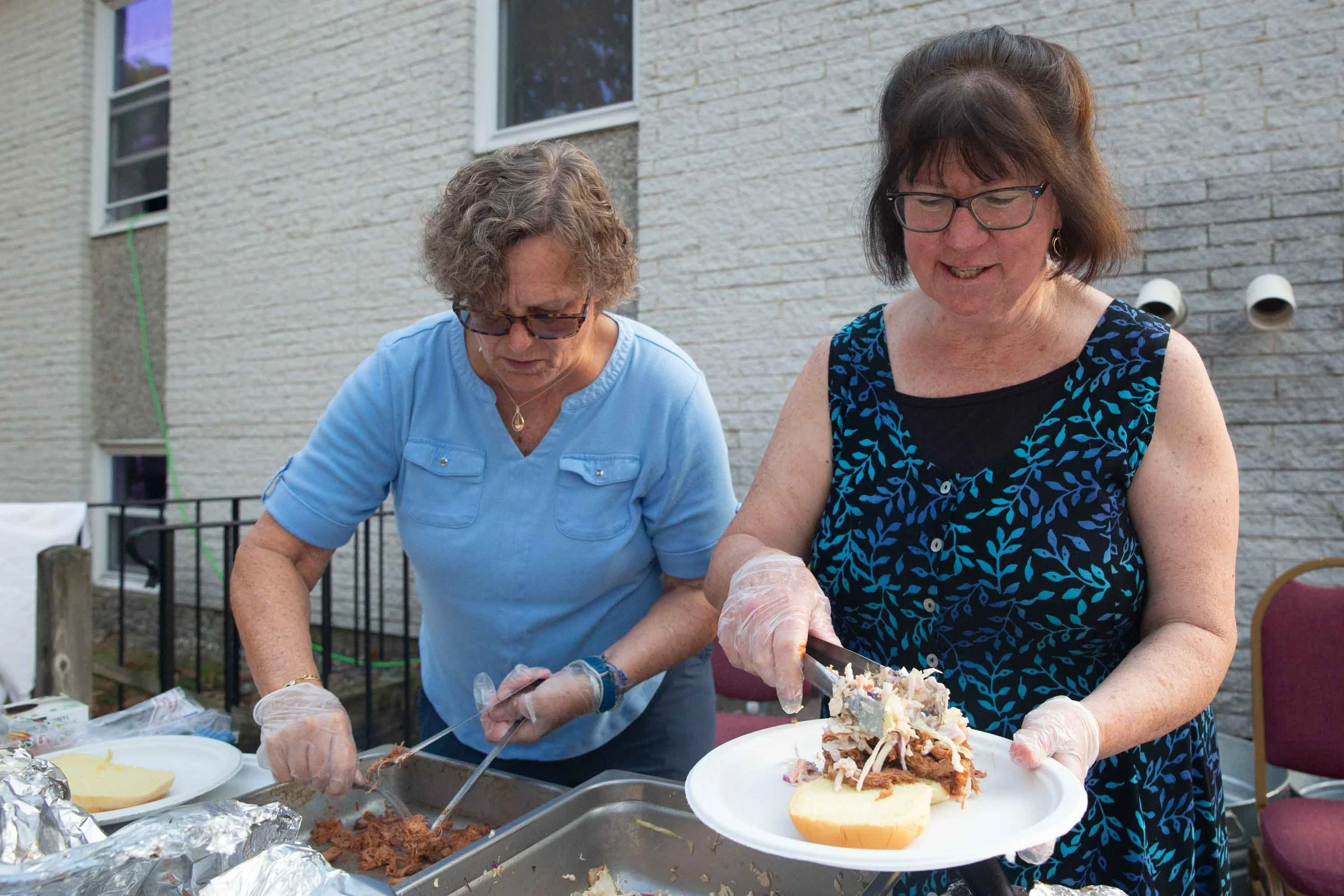 Two women preparing food outdoors, one is placing sliced potato on a plate and the other is serving shredded meat onto a plate. They are wearing plastic gloves and are standing in front of a table with food containers, in a backyard with a brick house wall and a railing.