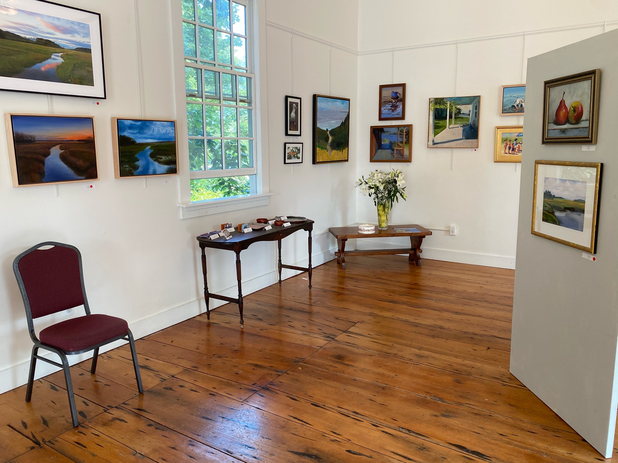 An art gallery with several landscape and still life paintings on white walls, a large window with natural light, a vase of white flowers on a small wooden table, and a maroon chair.