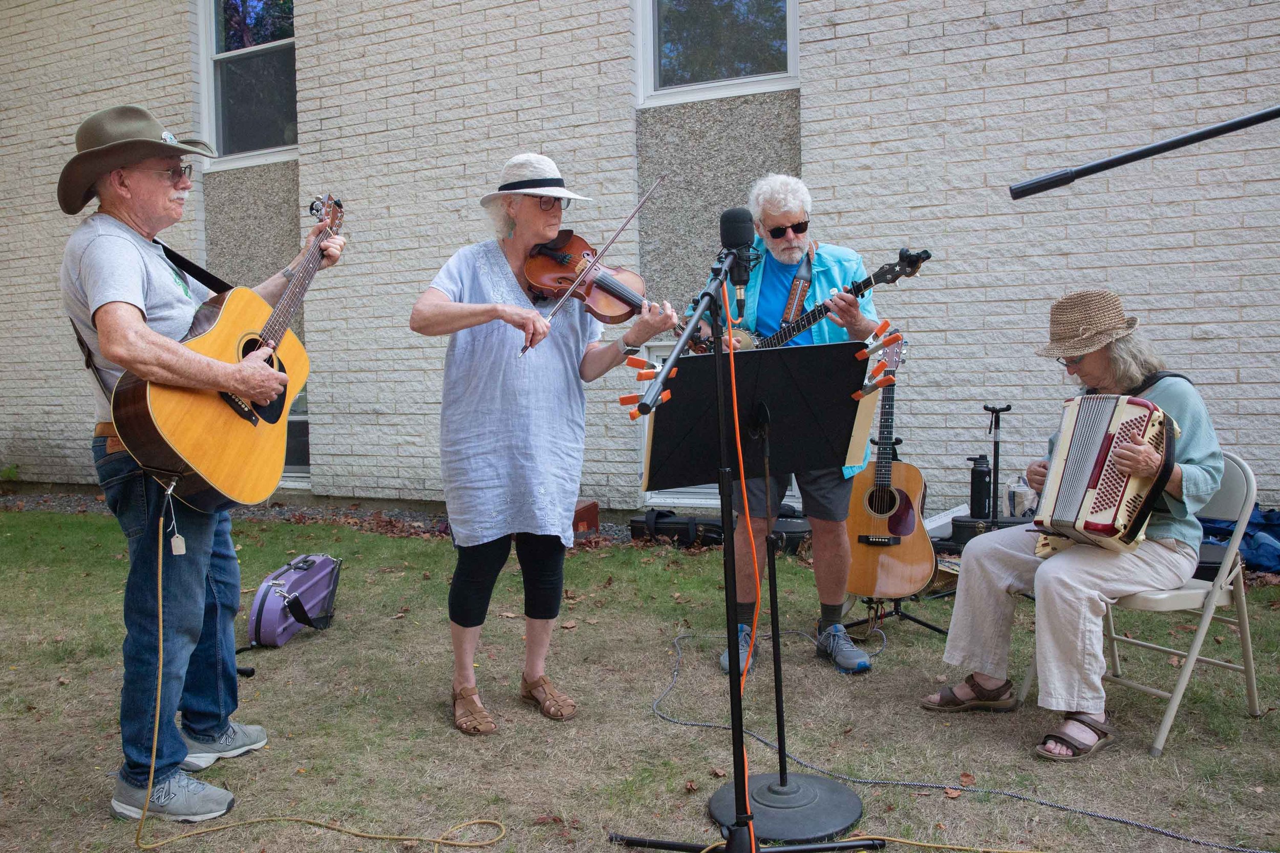 A group of five older adults performing music outdoors in front of a brick building. They are playing guitars, violin, and an accordion, with microphones and music stands set up.
