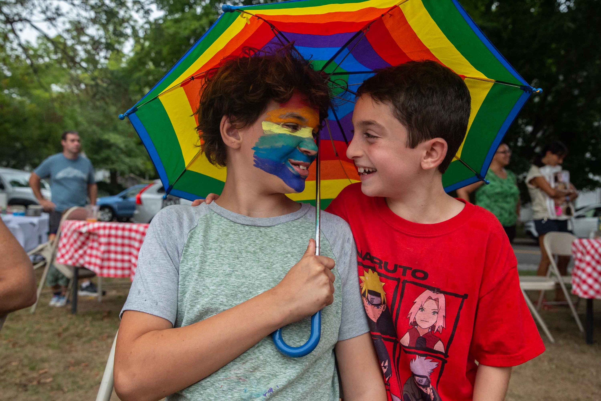 Two boys standing close under a rainbow-colored umbrella, with face paint on one boy, at an outdoor event.