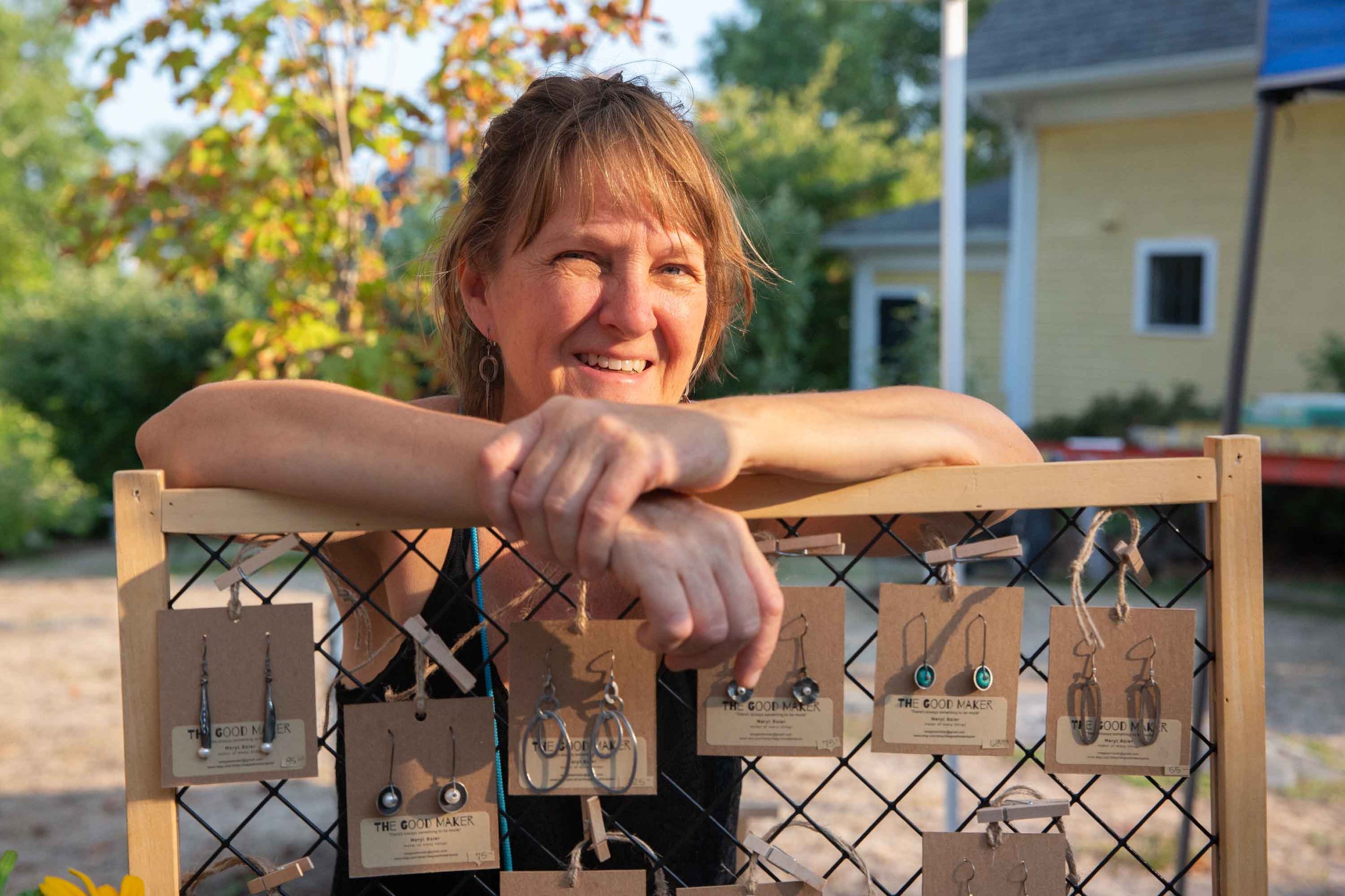 A smiling woman with short light brown hair leaning on a display rack of jewelry, including earrings, in an outdoor setting with trees and a yellow house in the background.