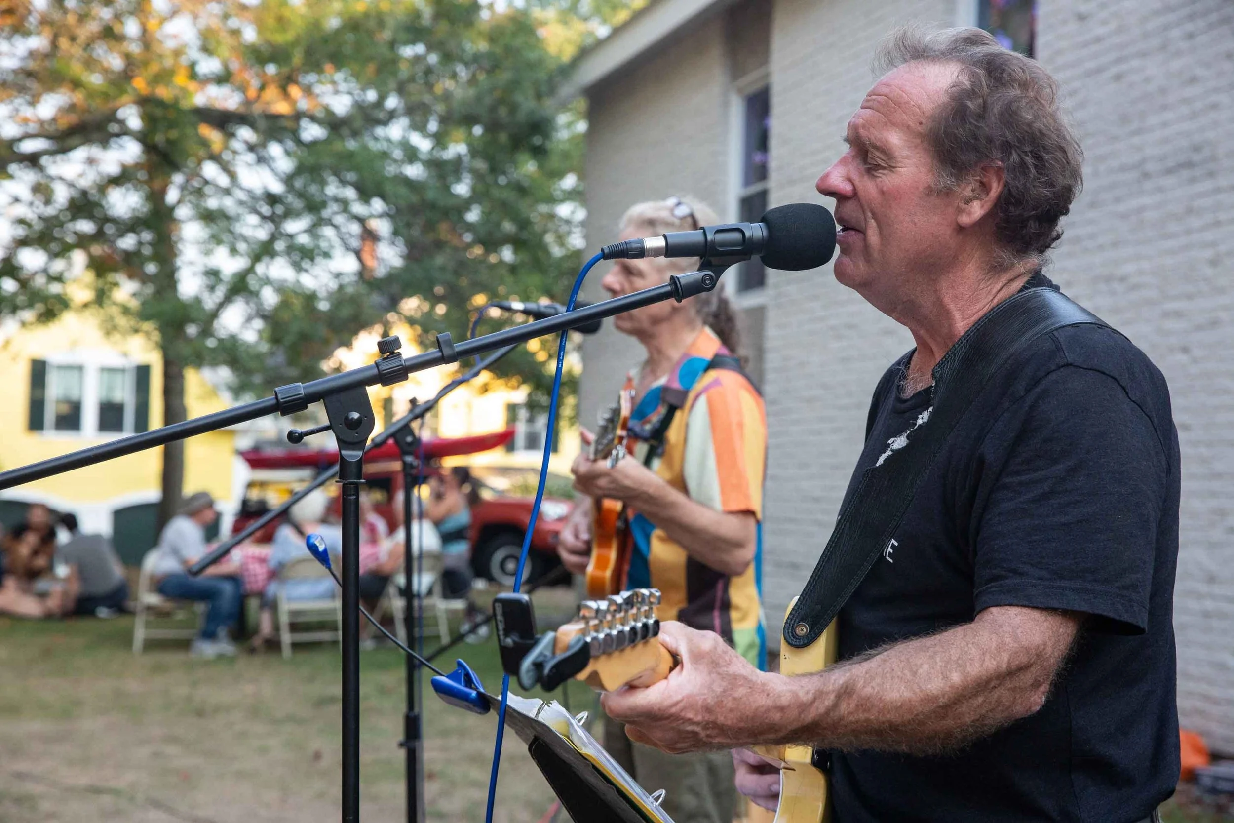 A man with curly hair singing into a microphone at an outdoor event, with a woman playing guitar behind him and multiple seated people in the background.