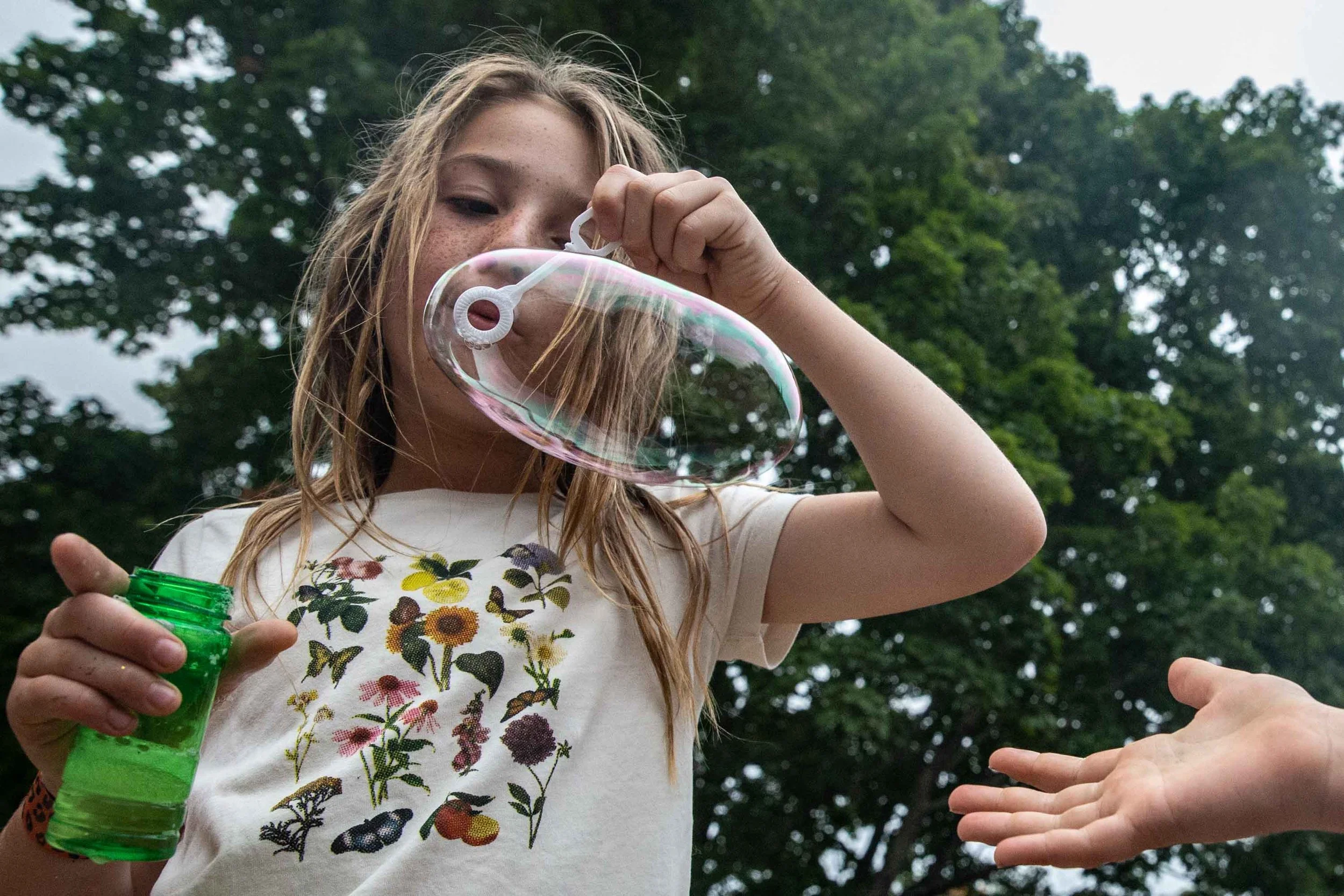 A girl with long blonde hair, freckles, and a floral t-shirt blowing soap bubbles outdoors. She is holding a green bubble solution bottle while another hand reaches in from the right side of the image.
