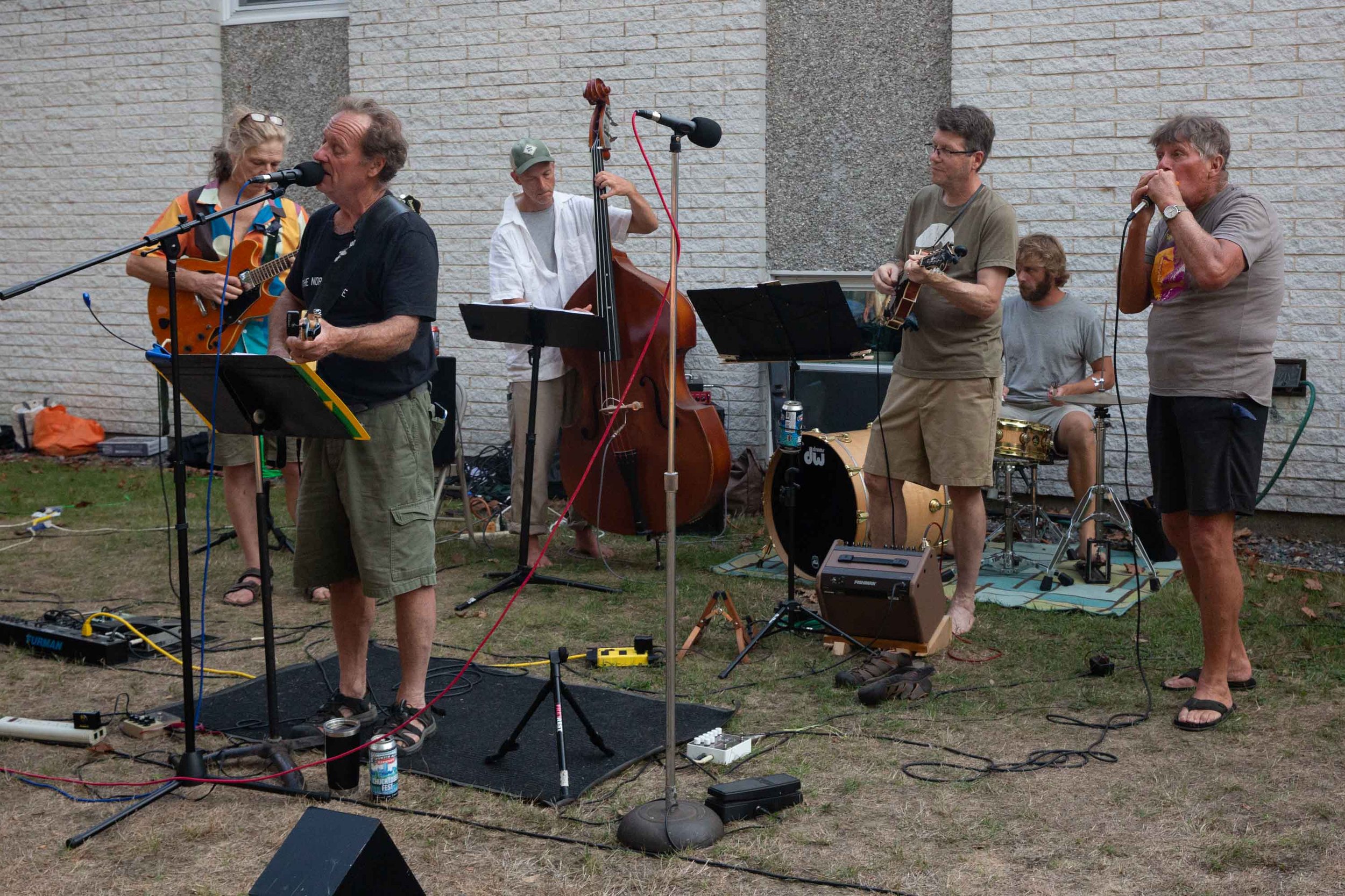A group of five men performing music outdoors in front of a brick wall. They are playing guitars, a double bass, and singing into microphones. There are various musical equipment and cables on the ground.