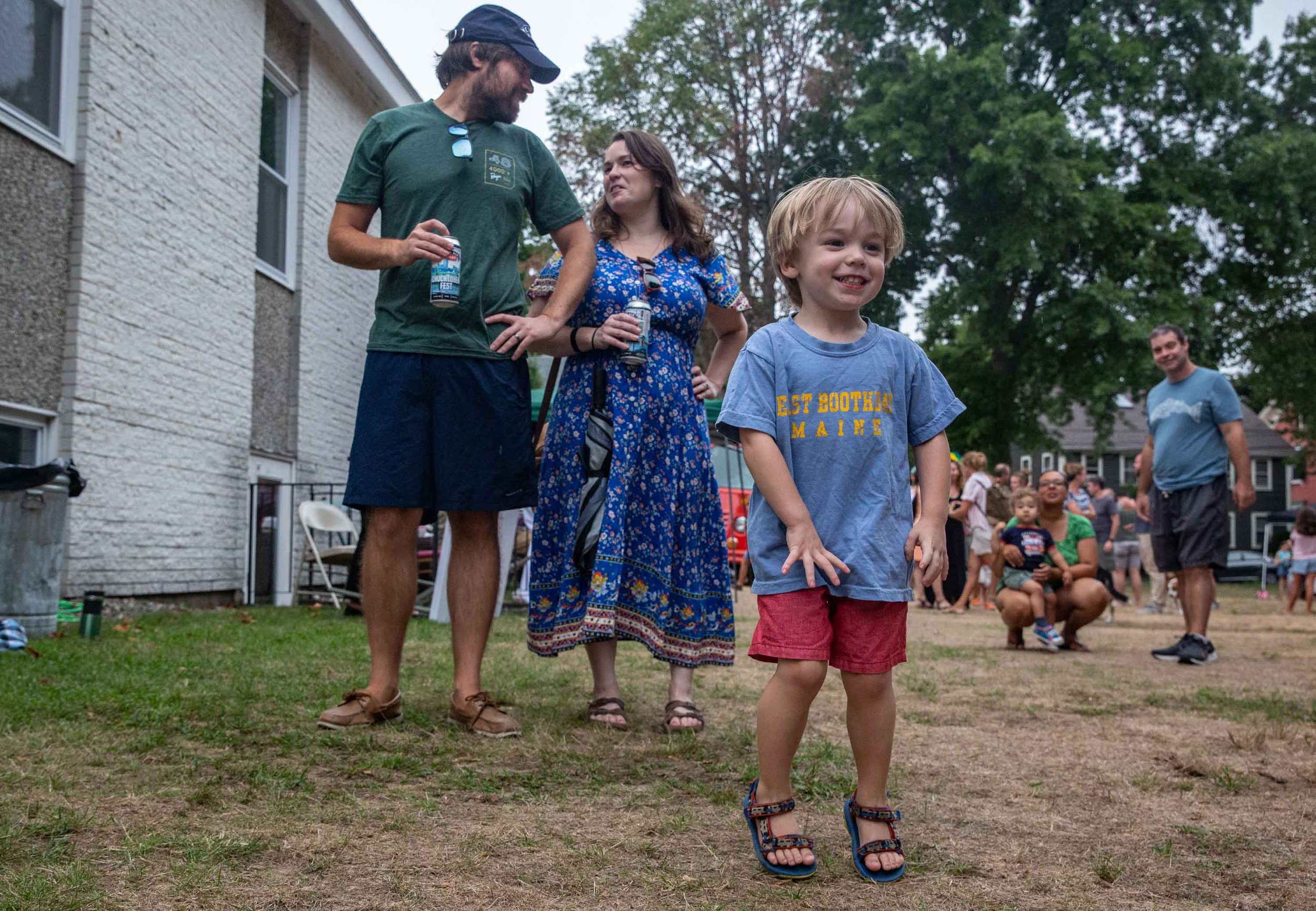 A group of people outdoors at a gathering, with a young boy in the foreground smiling, wearing a blue t-shirt and red shorts. Behind him, a woman in a blue floral dress and a man in a dark green t-shirt and cap are engaged in conversation. Several other people are in the background, sitting or standing, with trees and houses visible.