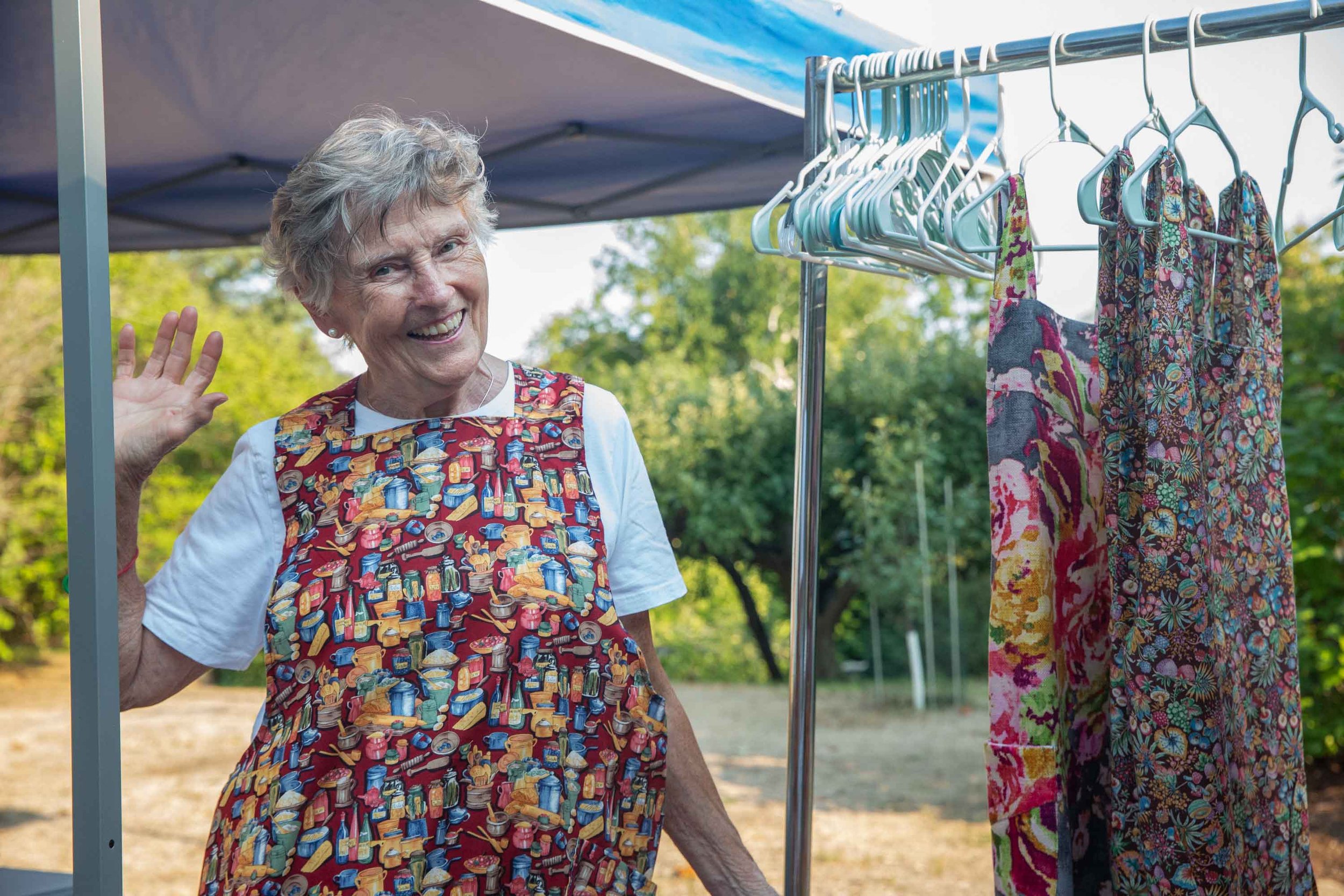 An elderly woman smiling and waving at an outdoor market stall with colorful clothing hanging on a rack, wearing a apron with a print of kitchen items.