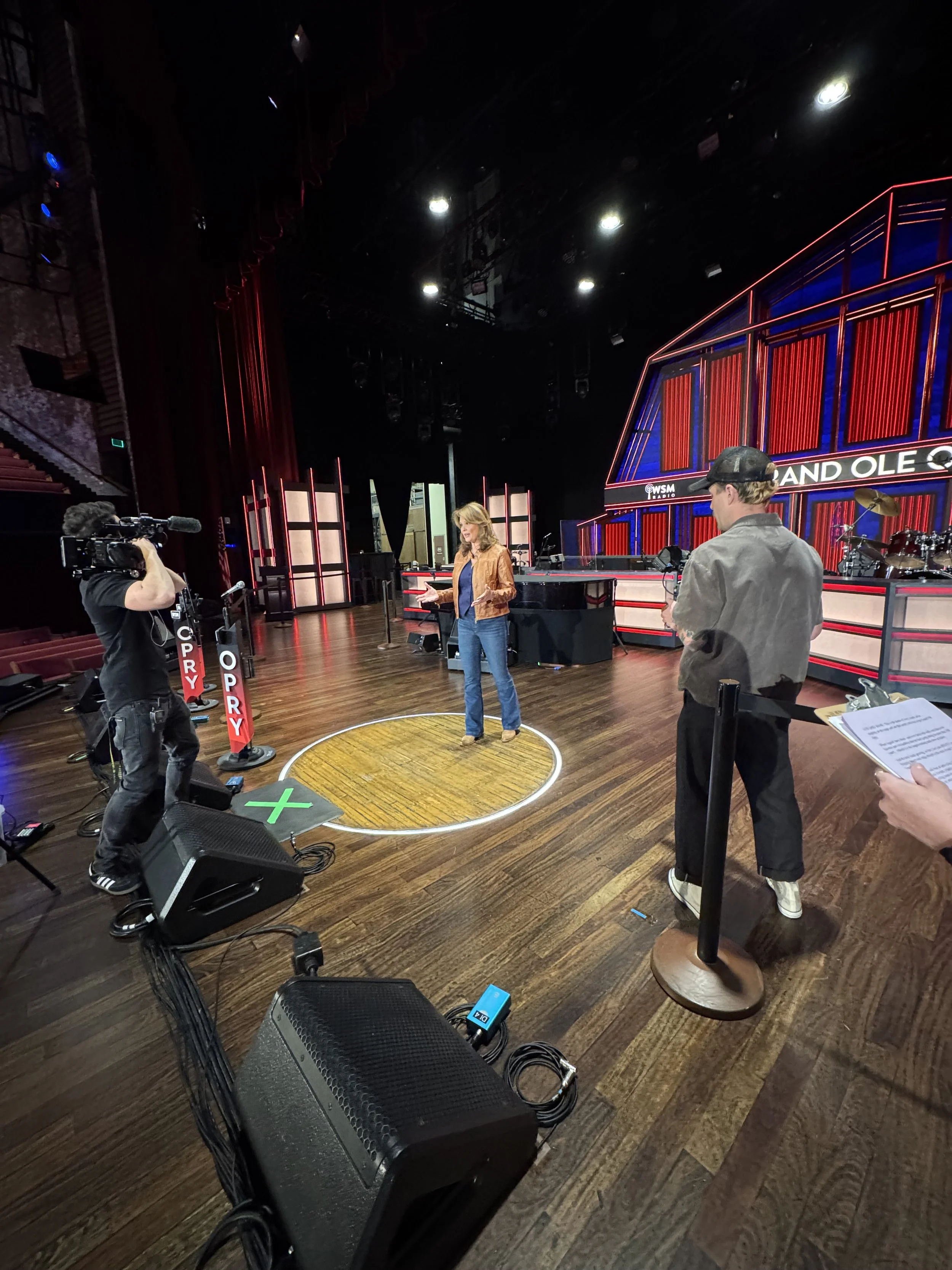 A woman is standing on a circular yellow rug on a wooden stage, talking with her hands raised. There are two men, one with a camera filming her and another with a clipboard. The background has a stage set with red and blue lights and the words 'LAND OLE' visible. The scene appears to be a television studio or stage setup.