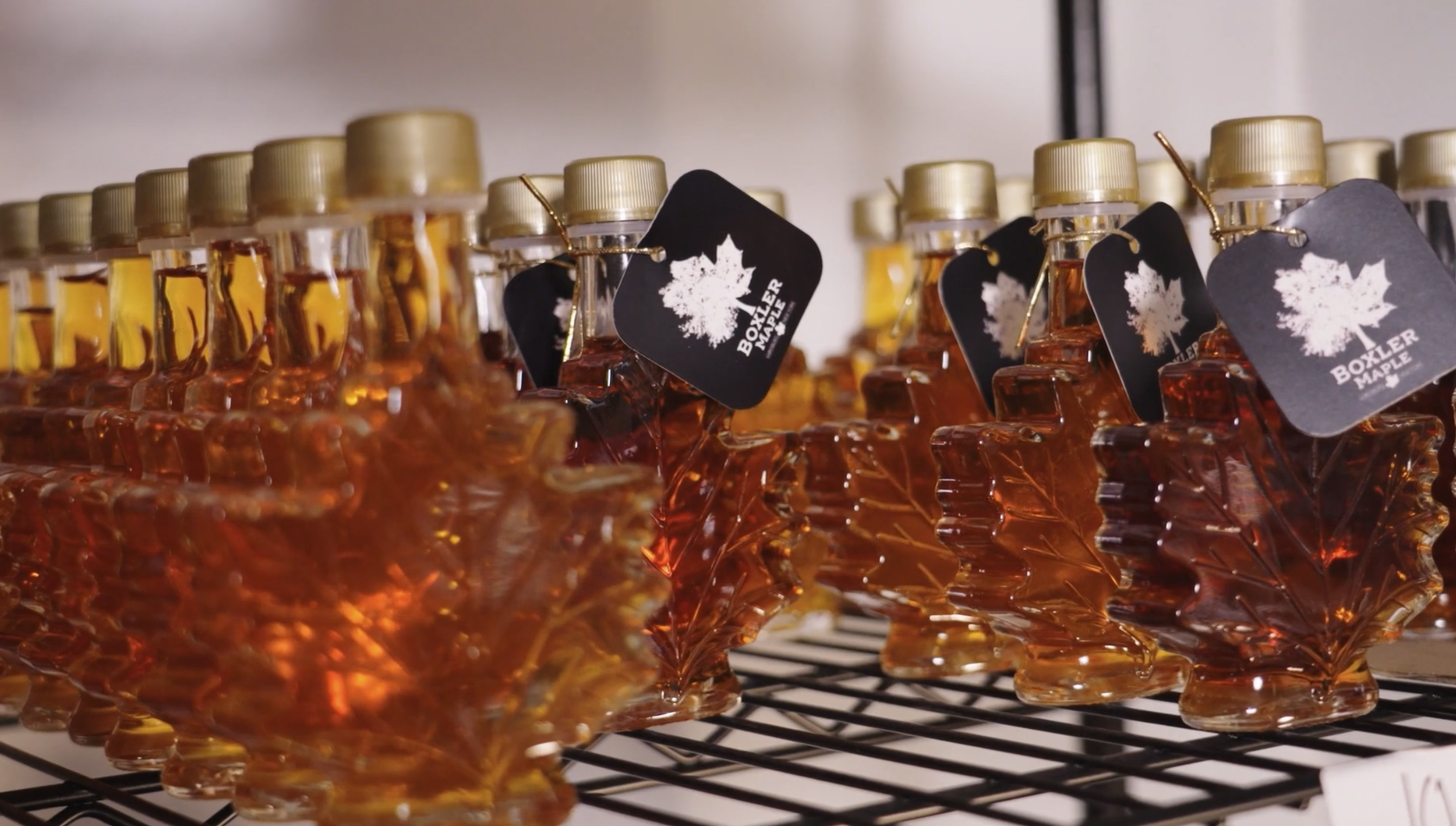 Rows of small glass bottles filled with amber-colored liquid, each with a black tag featuring a white maple leaf and the words "Boxler Maple," placed on a wire cooling rack.