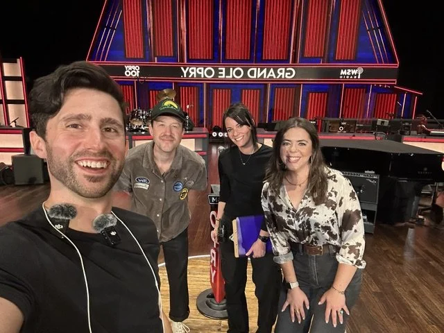 Four people taking a selfie in front of the set of the TV show "The Daily Show" with the "Grand Ole Opry" sign. The group is smiling and standing on a wooden stage with various equipment around them.