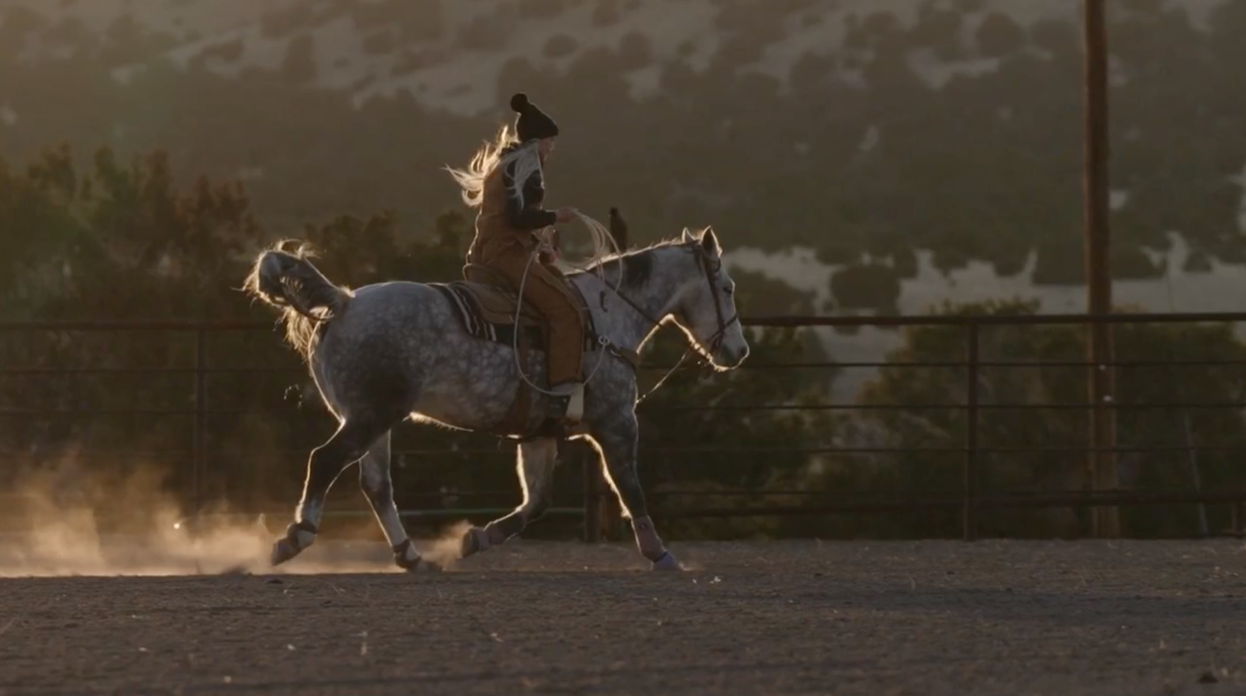 A woman riding a gray horse with white spots in an arena, wearing a winter hat and casual riding clothes, during late afternoon or early evening.