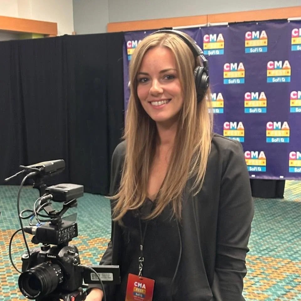 A woman at a media booth wearing headphones, smiling, with a camera on a tripod, and a background with 'CMA' and 'SoFi' logos.
