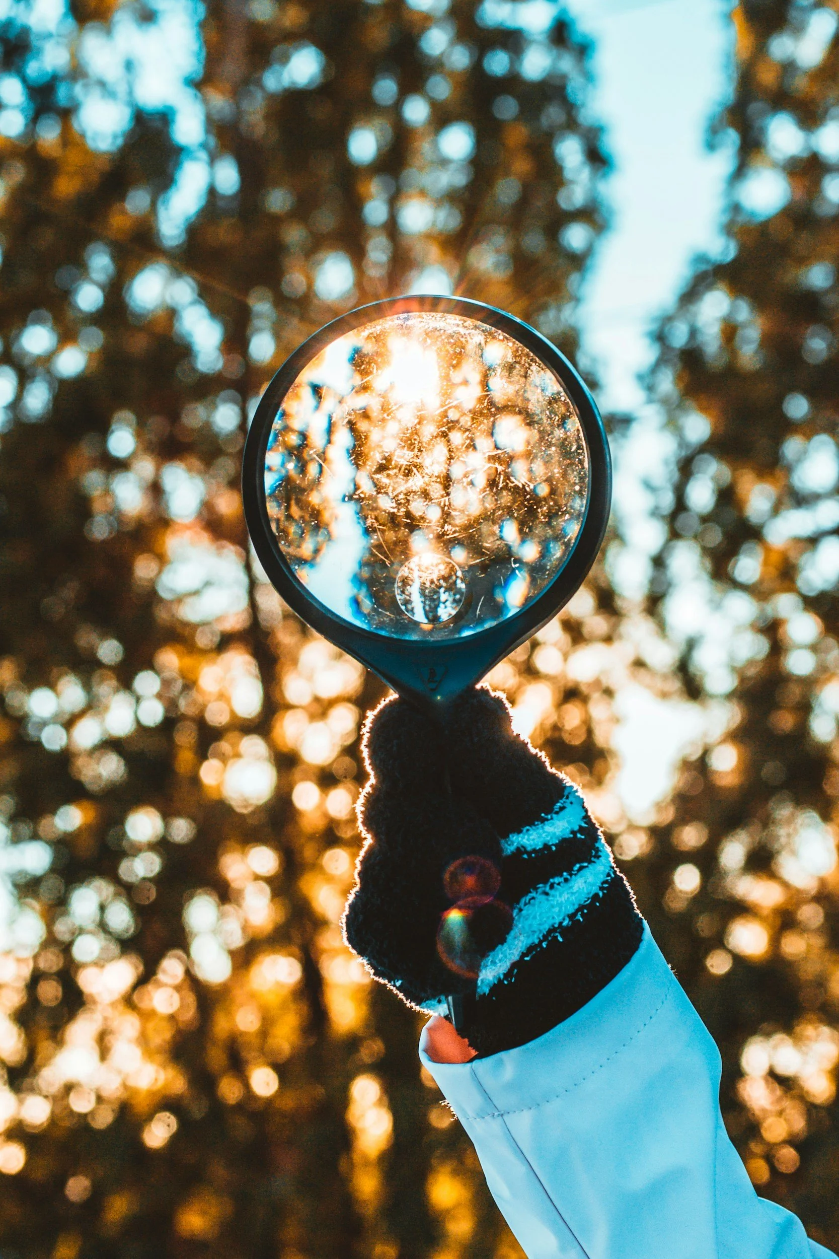 A person wearing a black glove with white stripes, a white jacket, and holding a reflective lens up to the sun. The sun shines through the lens, creating a bright light and reflections, with background of blurred trees and clear sky.