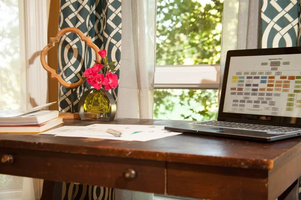 A wooden desk with a laptop, some papers, and a pair of glasses on it, positioned in front of a window with green trees outside. There is also a small vase with pink flowers and a decorative wooden mirror on the desk.