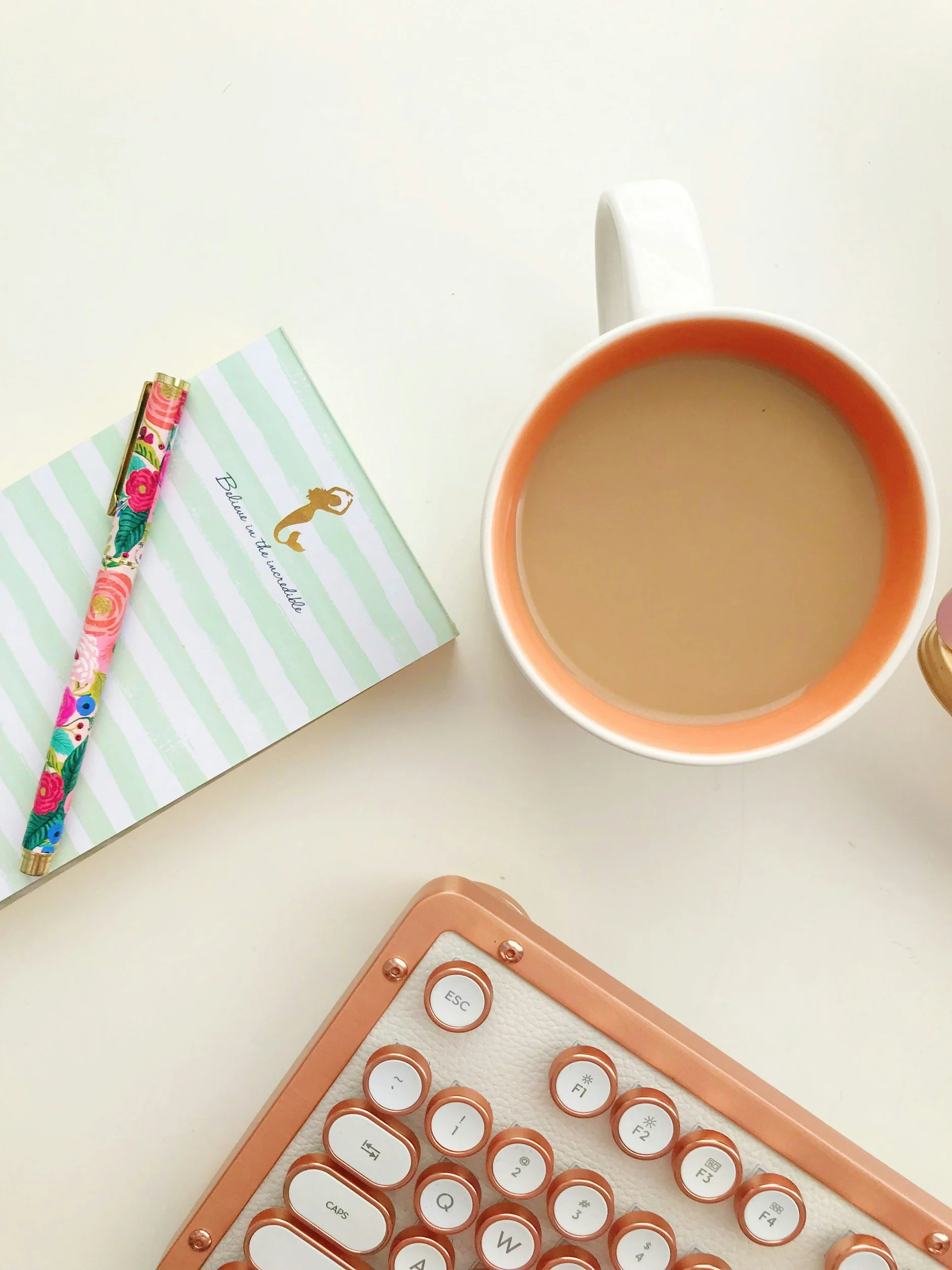 Top view of a desk showing a pink keyboard, a light green and white striped notebook, a floral pen, a white mug filled with coffee or tea, and part of a pink electronic device.