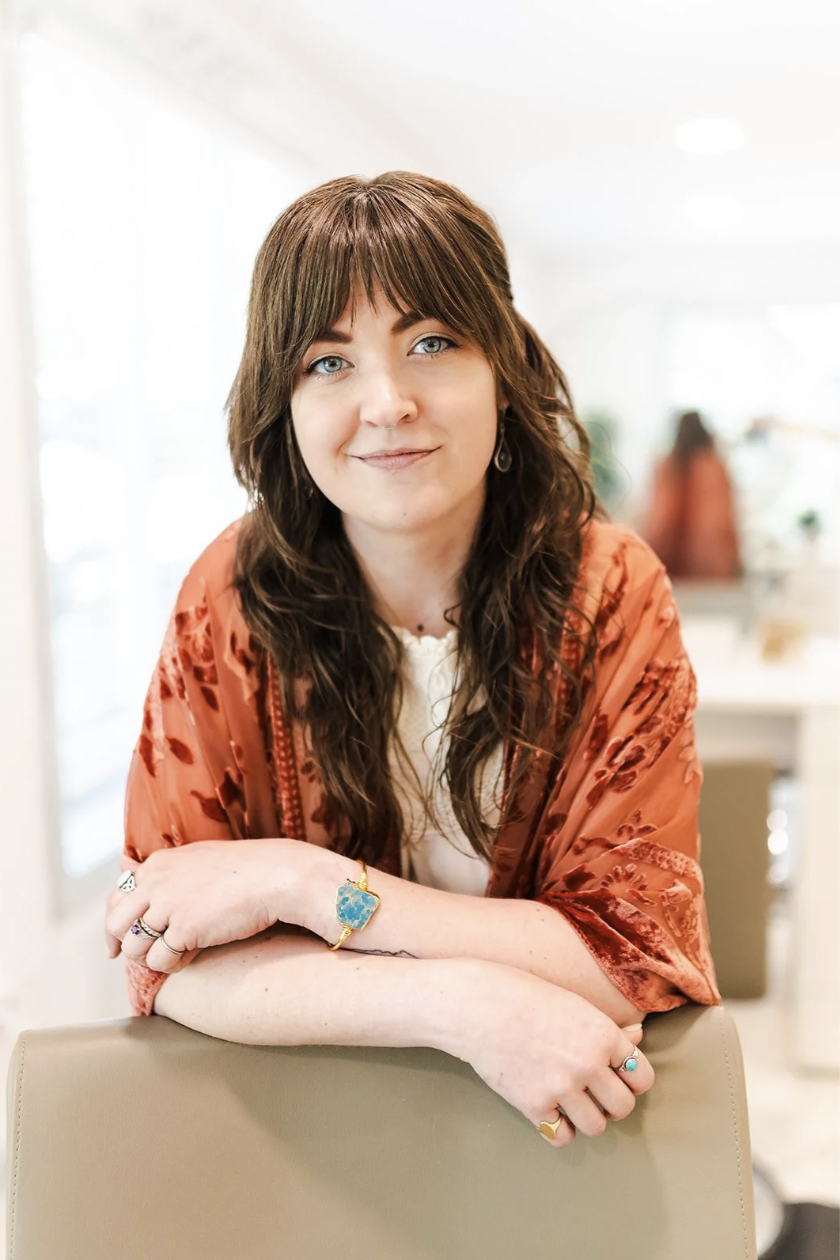 Holly Millar White, haircutting specialist, with long wavy brown hair and bangs, leaning on a salon chair