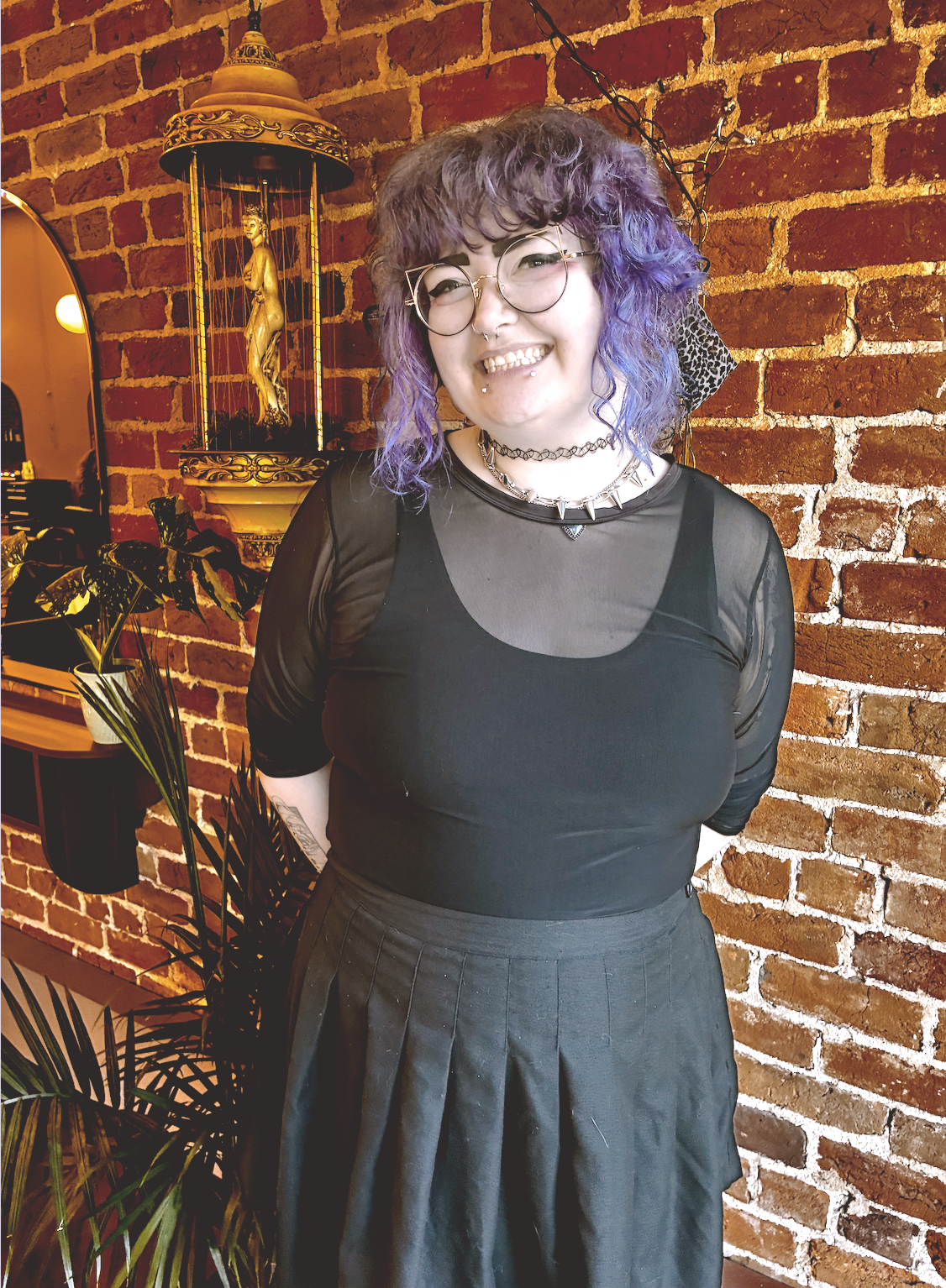 Max Balcom, haircutting specialist, with curly violet hair and round glasses against the brick wall at The Darling