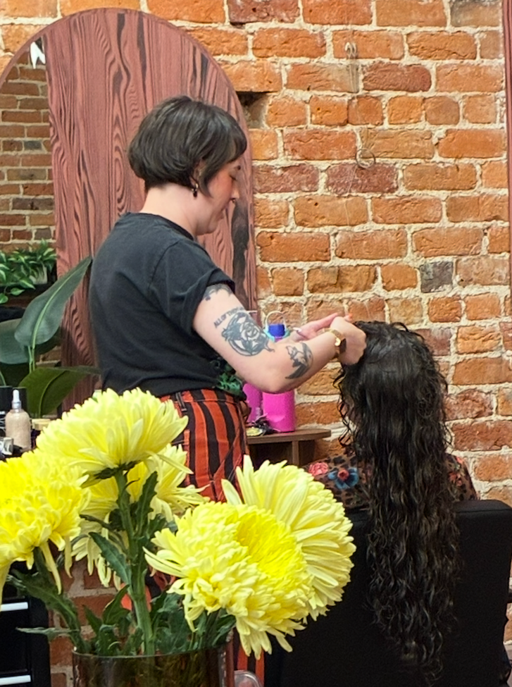 Hairstylist cutting a client's long curly hair in a salon with yellow flowers and exposed brick walls