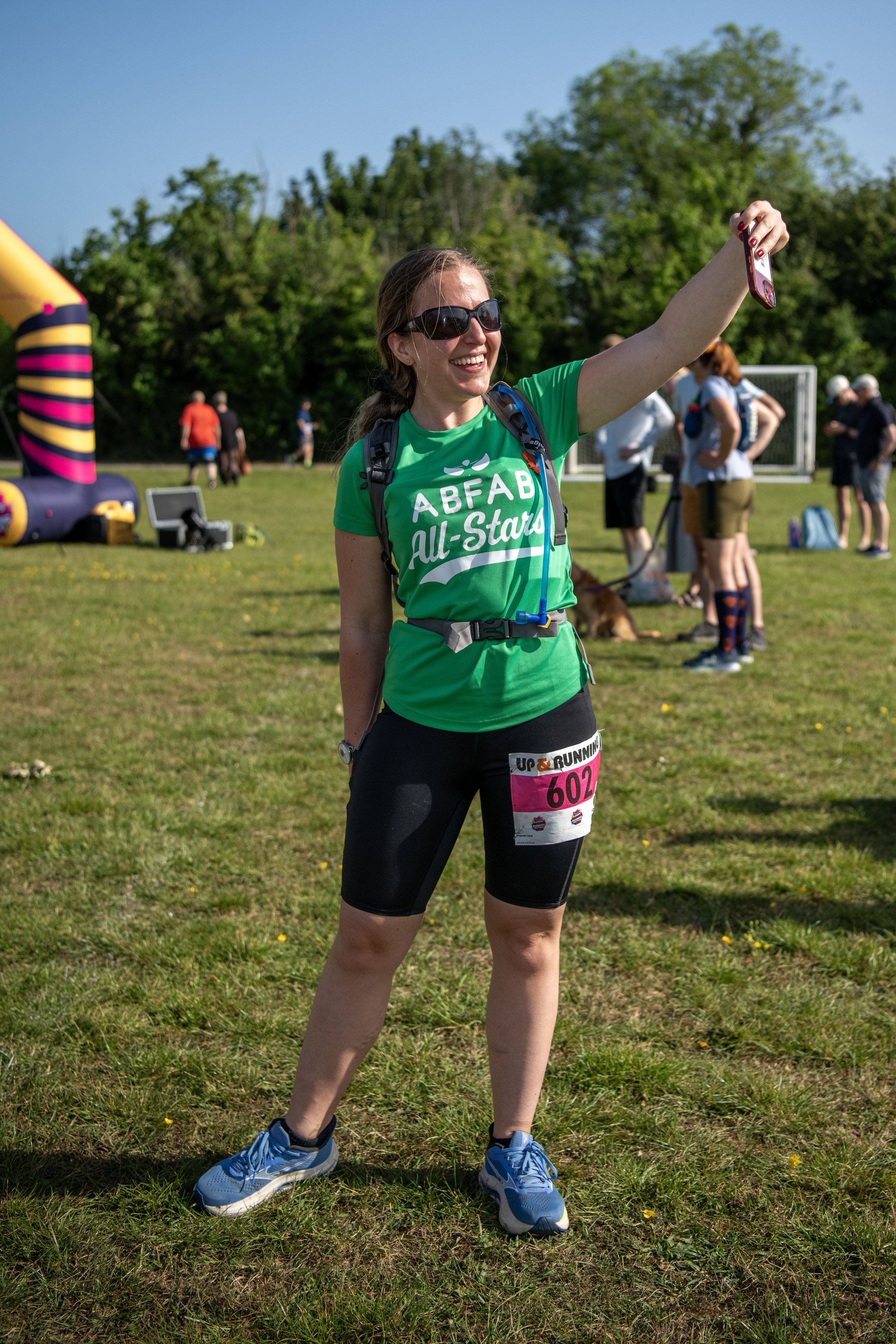 A woman wearing sunglasses, a green shirt with 'ABFAB All-Stars' written on it, black shorts with a pink race bib numbered 602, and running shoes, taking a selfie at an outdoor event on a sunny day. There are other people and a soccer goal visible in the background.