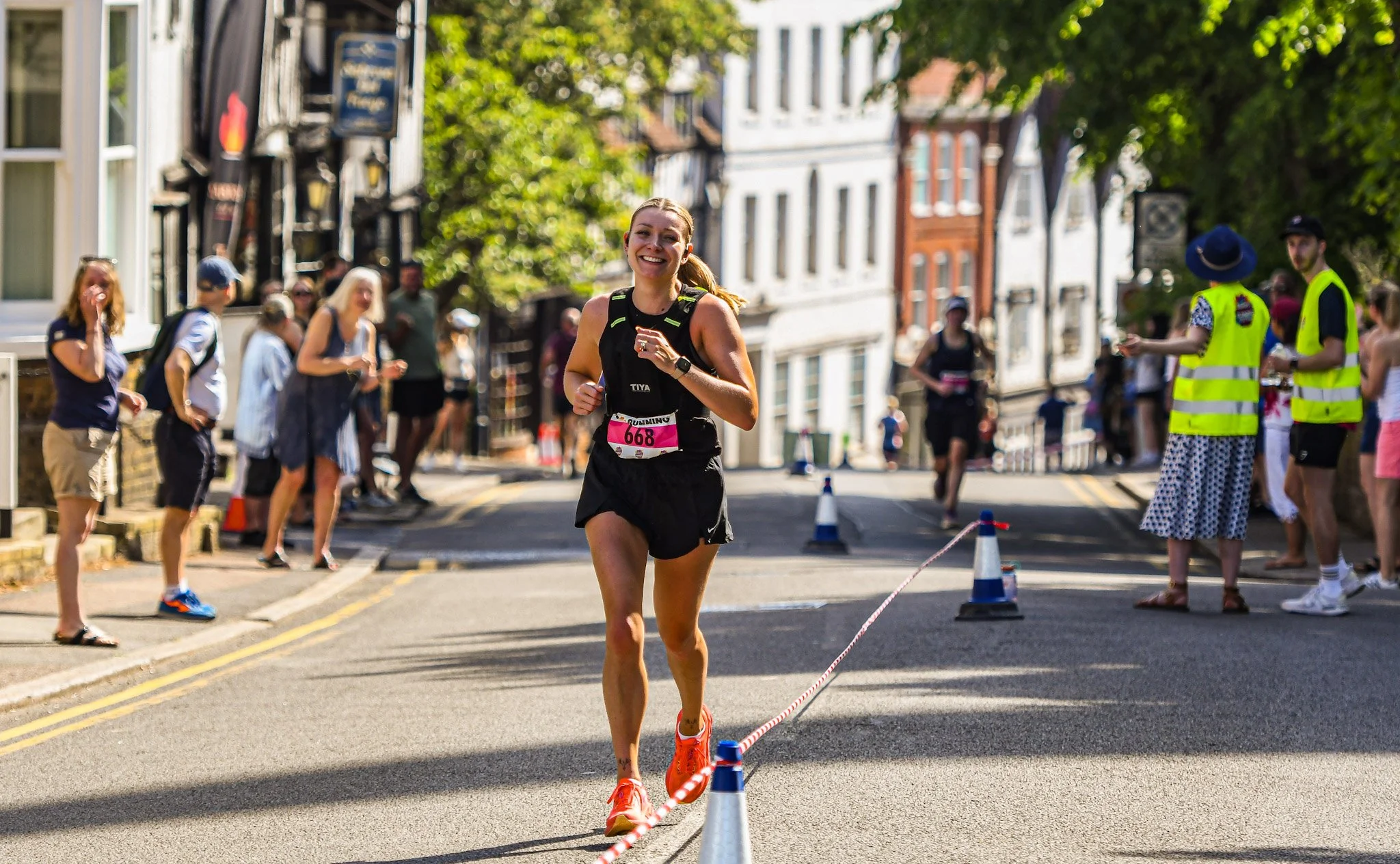 A woman running in a race during daytime, smiling, wearing a black tank top and shorts, with a pink race bib number 668. Spectators and race officials are seen on the sides of the street, some taking photos and others cheering. The street is lined with trees and historic buildings.