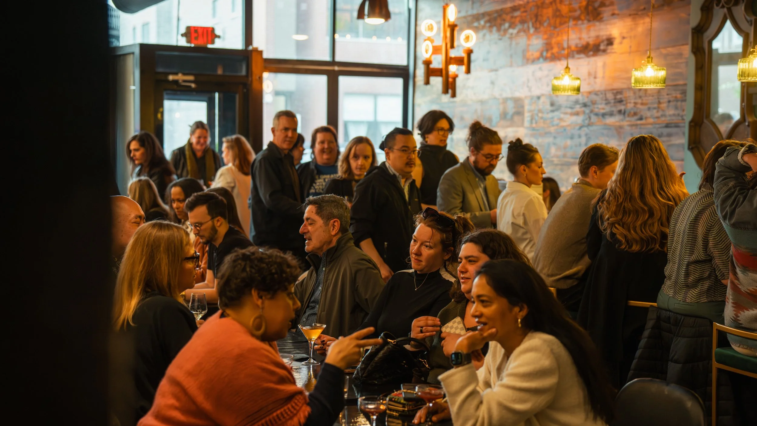 People socializing and chatting at a busy restaurant or bar with warm lighting, brick wall, and large windows.
