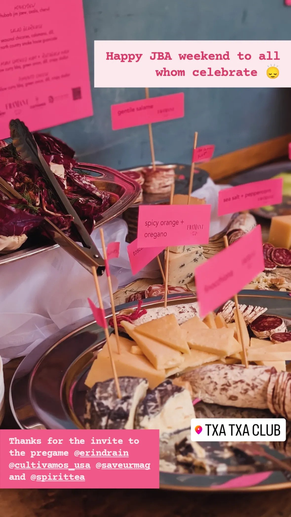 A cheese and charcuterie platter with assorted cheeses, cured meats, crackers, and pink labels indicating different food items, set on a table for a gathering or celebration.