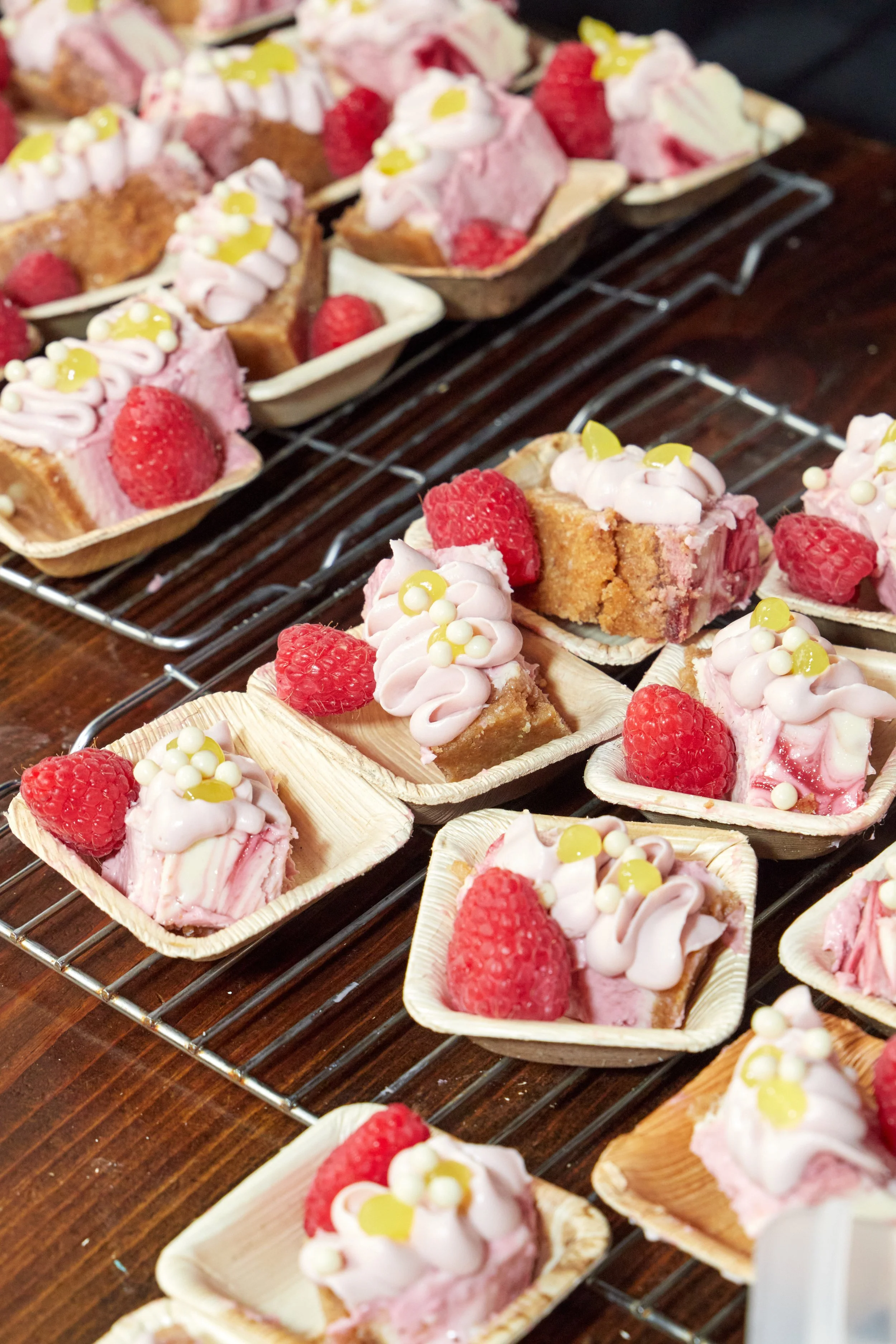 Various strawberry and raspberry desserts decorated with pink icing, white spheres, and yellow-green drops on individual cupcake liners on a wire cooling rack.