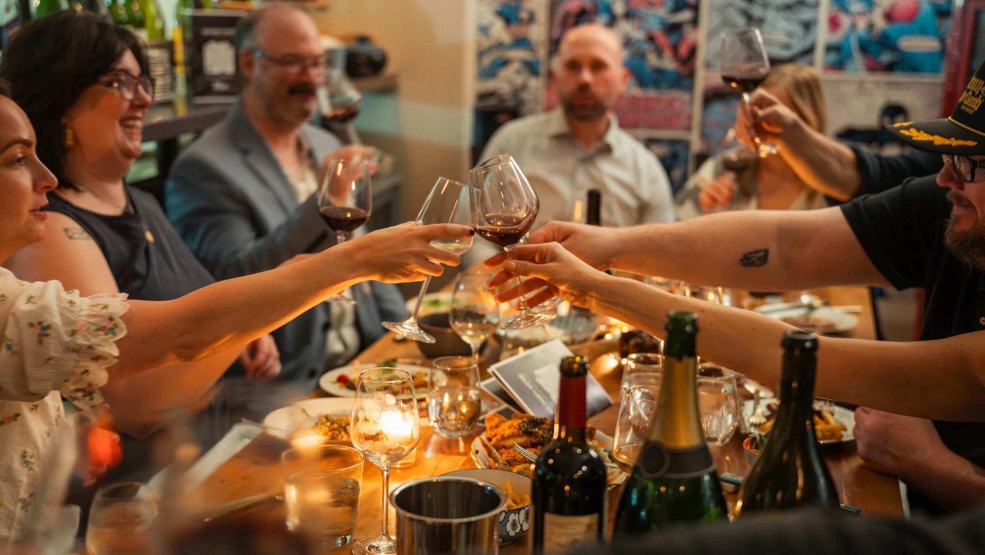 A group of people around a table at a dinner party, raising glasses of red wine in a toast, with food and drinks on the table.