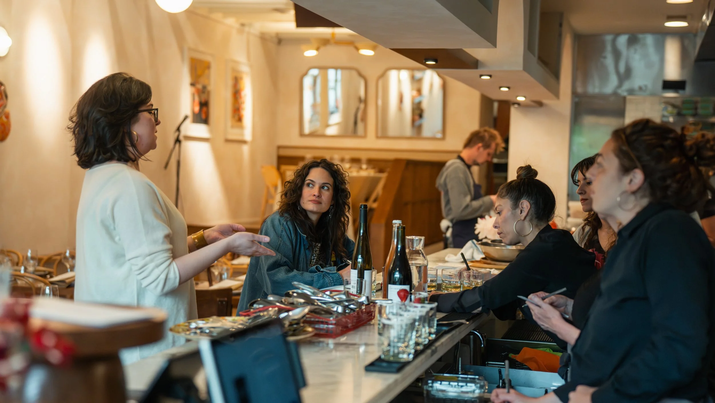 A woman standing and speaking to a group of women seated at a restaurant counter, with wine bottles and glasses on the counter.