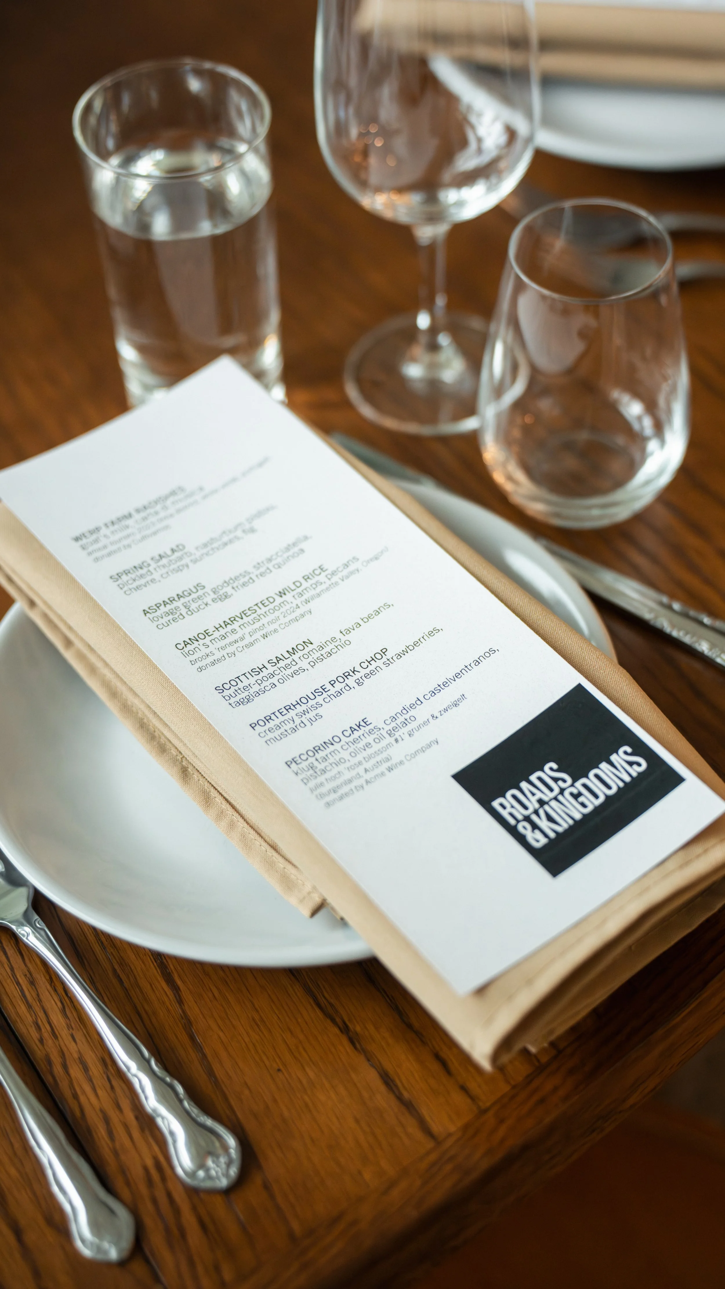 A restaurant table setting with a menu, a plate, silverware, a glass of water, and two empty wine glasses on a wooden table.