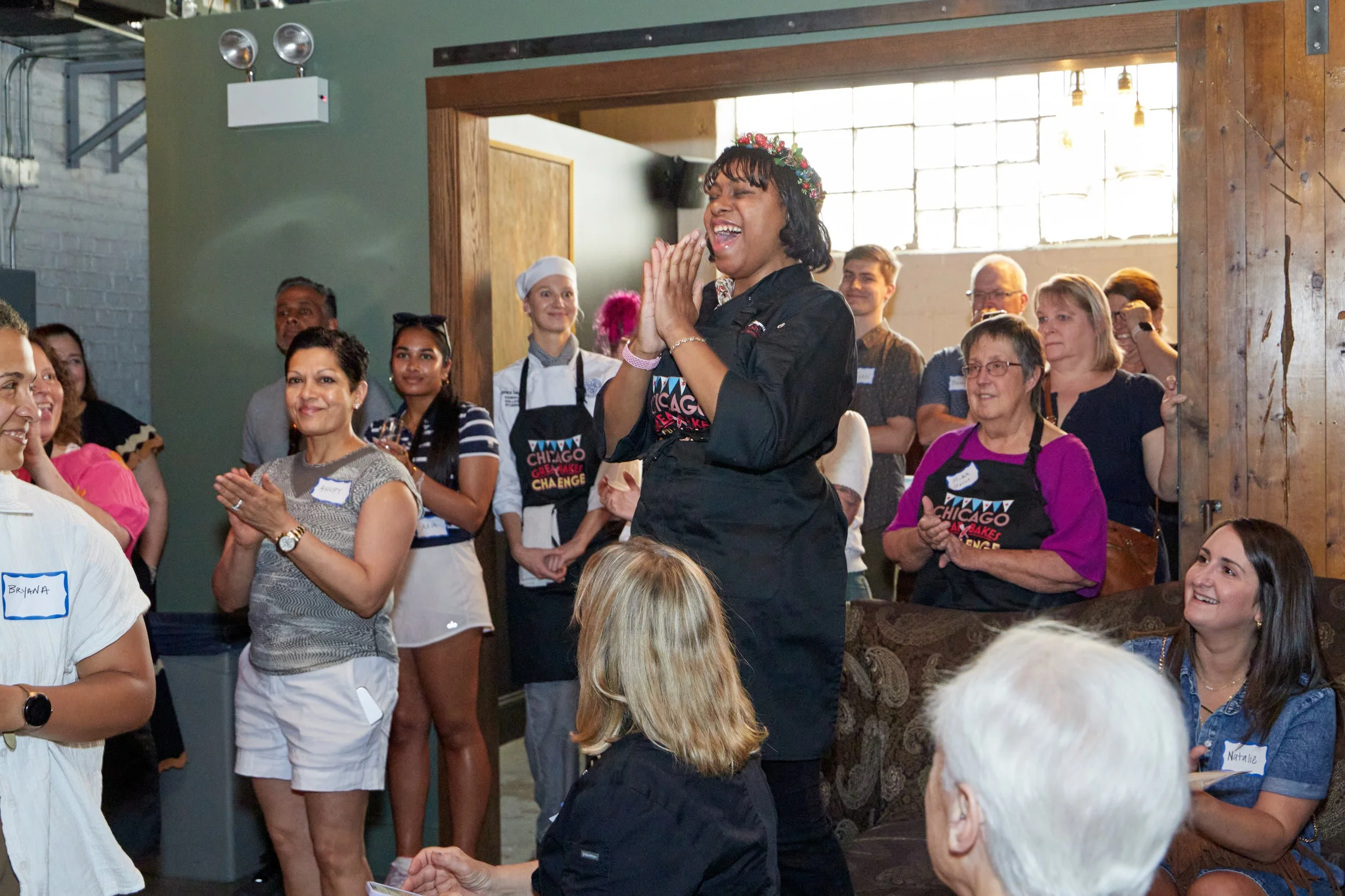 A woman with dark hair is smiling and clapping, surrounded by seated and standing people at an indoor event with natural light and exposed brick walls.