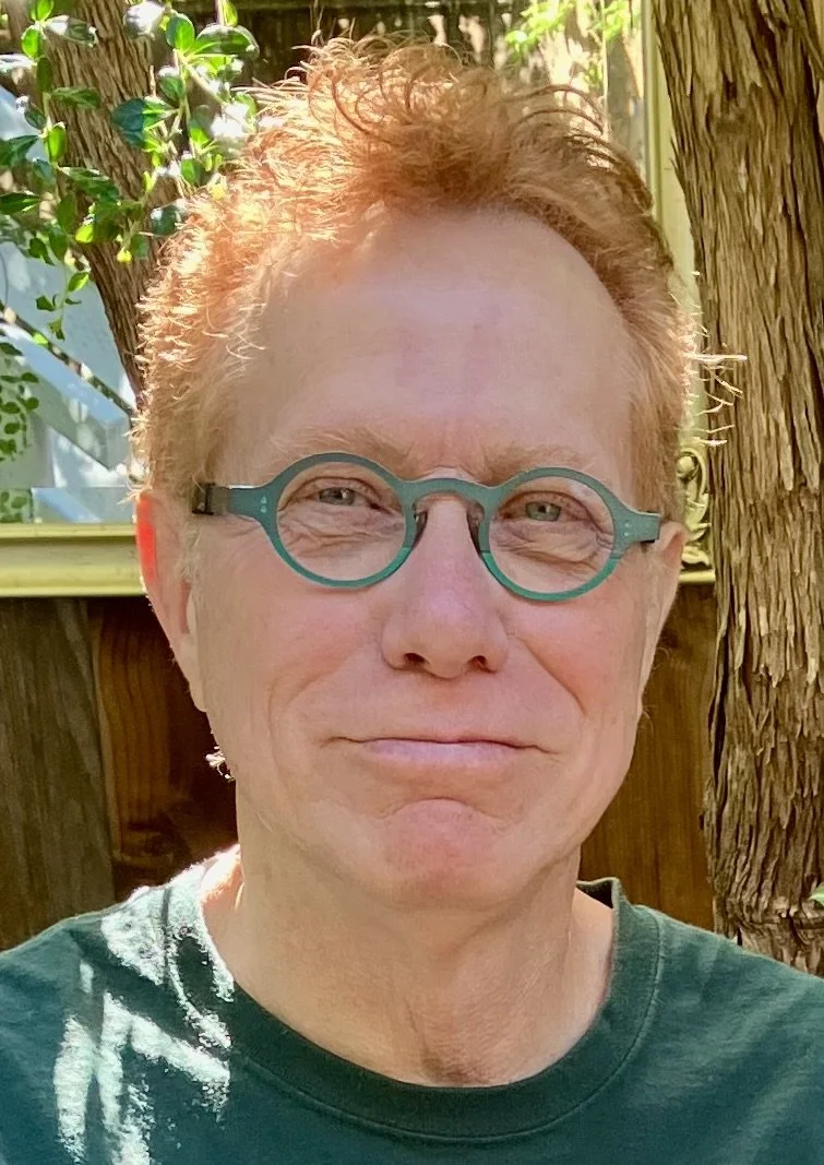 A person with short, curly reddish hair wearing teal round glasses, standing outdoors next to a tree with green leaves in the background.