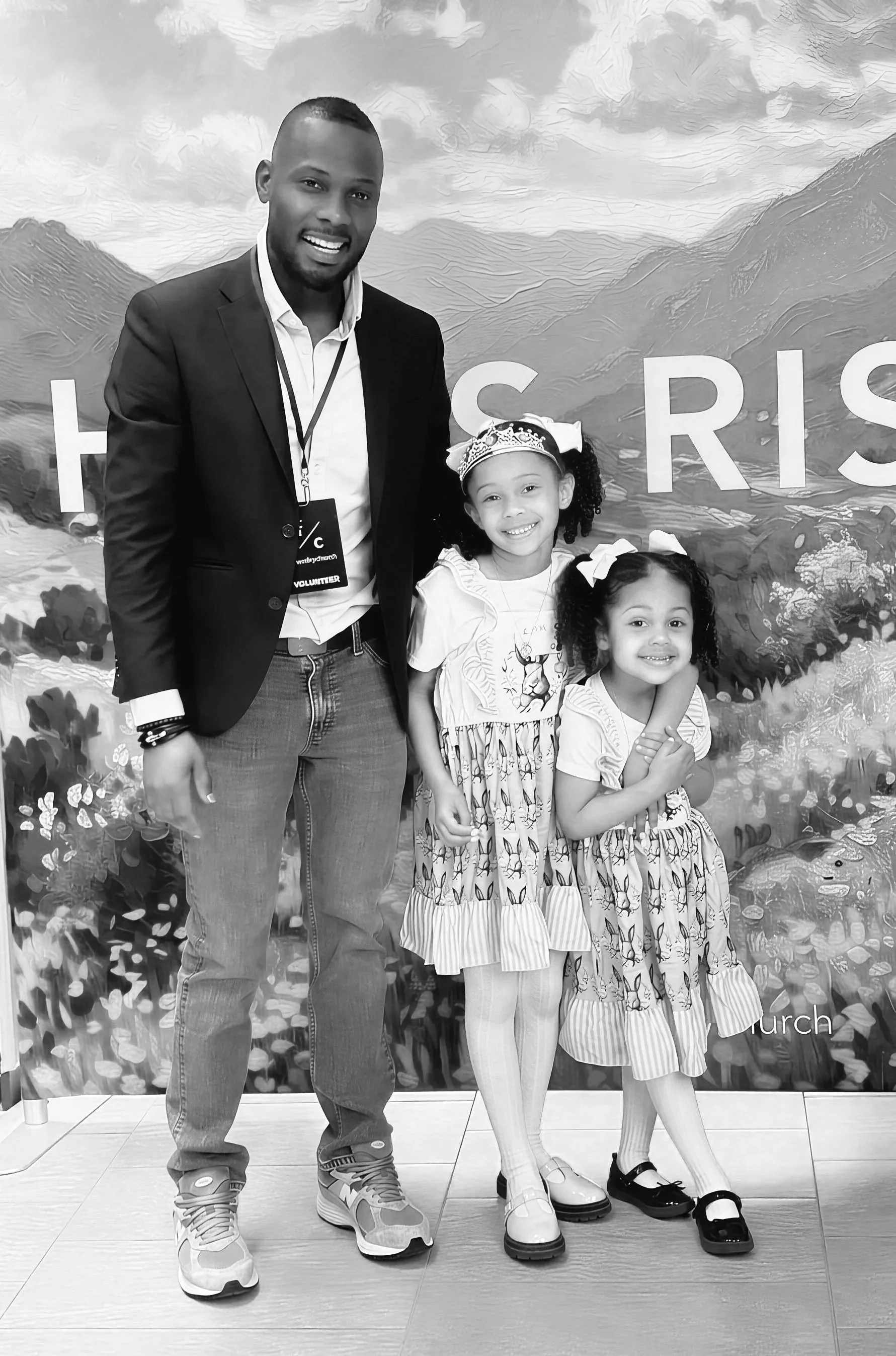 Four people, one adult man and three young girls, standing in front of a nature-themed backdrop with mountains and trees, smiling at the camera. The man is wearing a dark jacket,.t-shirt, jeans, and sneakers. The girls are in dresses with patterns and bows in their hair, with two of them wearing heels and one wearing black shoes.
