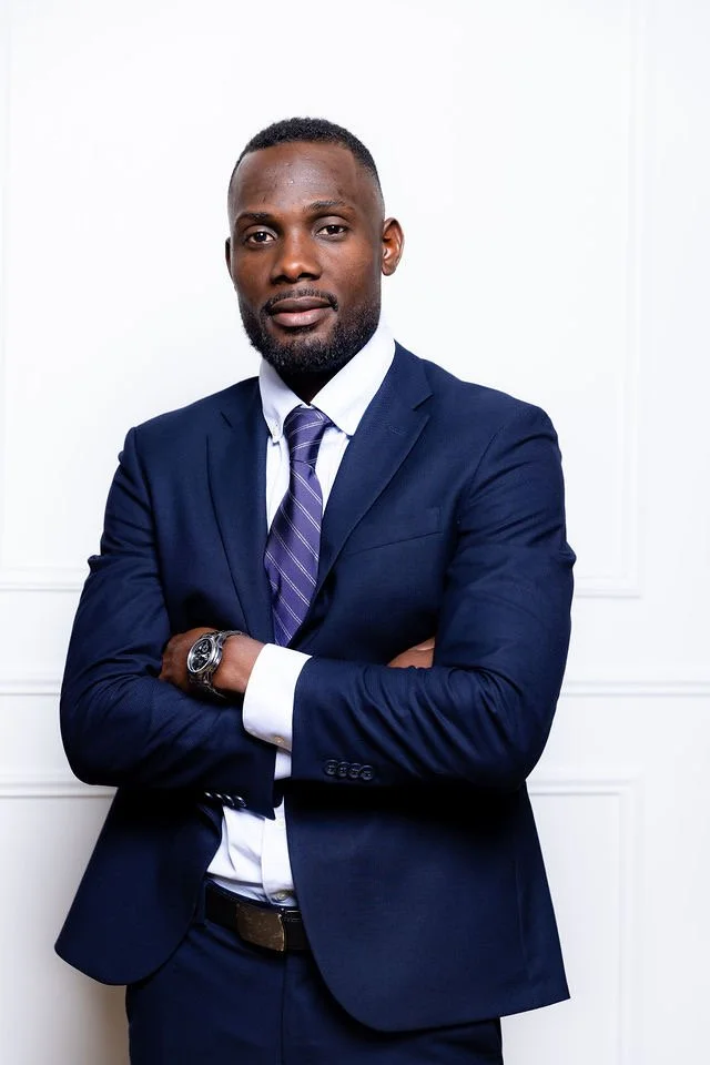 A confident African American man in a navy blue suit, white shirt, and purple tie stands with arms crossed against a white background.