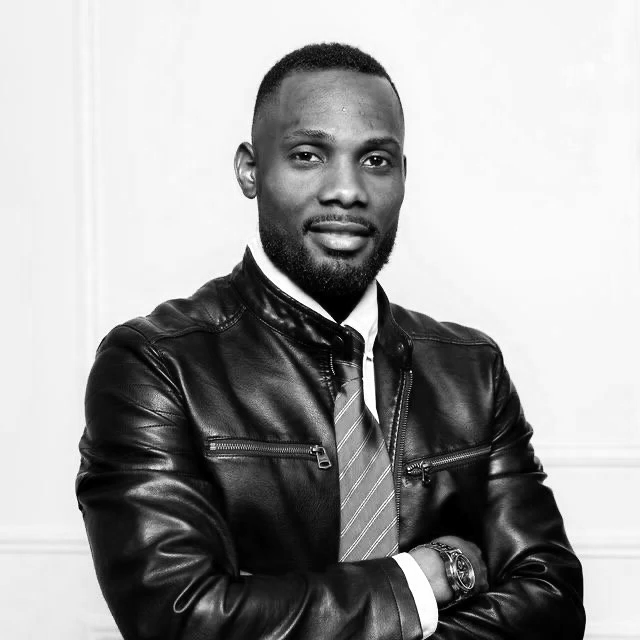 A Black man with a short beard wearing a leather jacket, striped tie, dress shirt, and watch, posing with arms crossed in front of a plain wall.