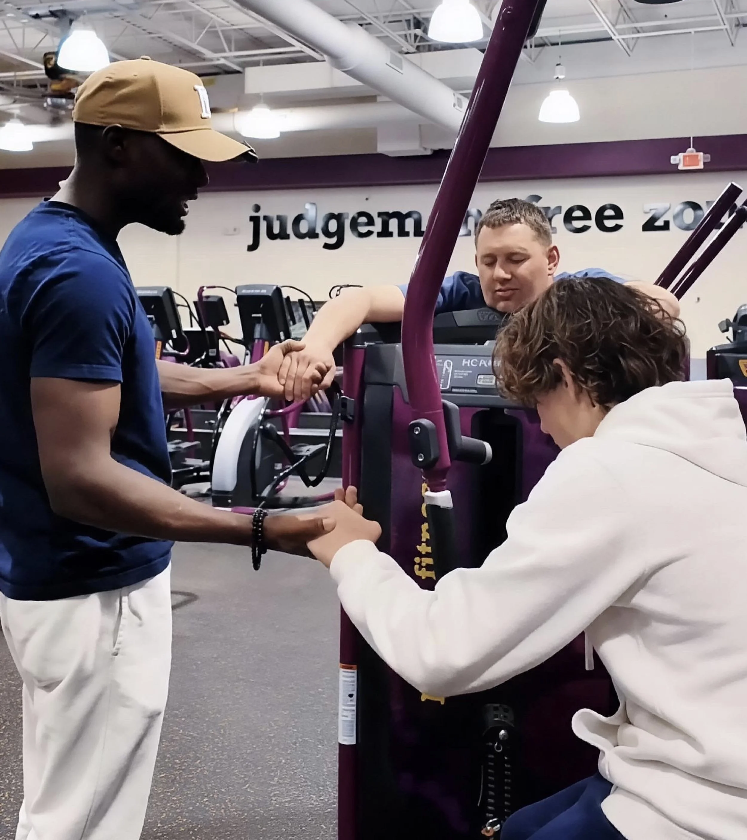 Group of young men in a gym or fitness center, with one man in white pants holding a gym equipment handle while another man in a beige baseball cap and blue shirt looks on, and a third man in a white hoodie also engaged in the activity.