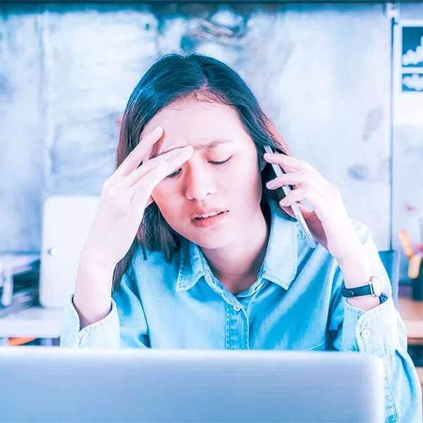 Woman experiencing headache at work, holding temples with closed eyes, sitting at desk with computer.