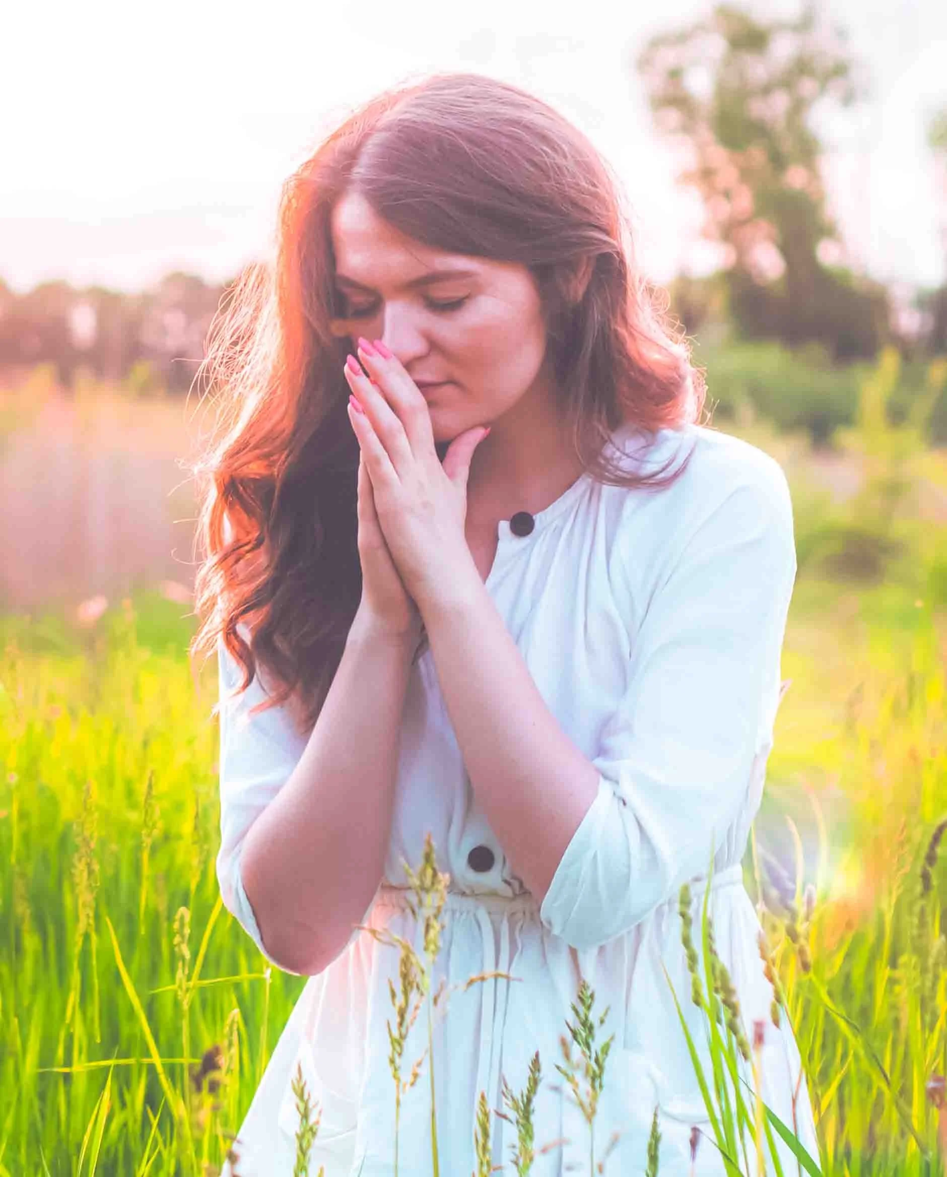 Young woman with long red hair standing in a green field or meadow, her hands pressed together near her face, with sunlight illuminating her hair from behind.