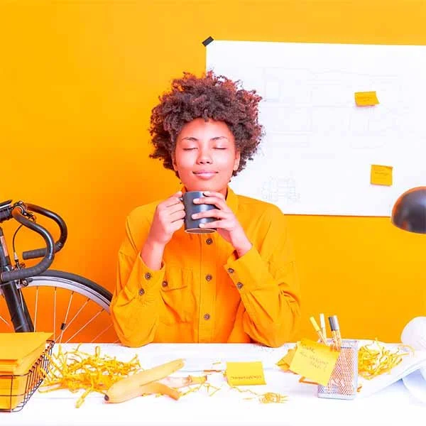 A woman with curly hair and a yellow shirt sitting at a desk with a cup in hands, eyes closed, smiling, in a bright yellow room with a whiteboard behind her.