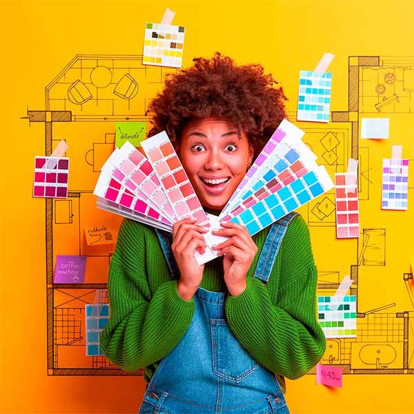 a woman with curly hair smiling, holding color swatches in front of a yellow wall withsketches and notes related to interior design