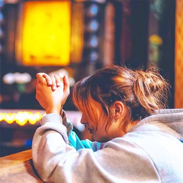 A woman with brown hair in a ponytail wearing a gray hoodie, resting her head on her forearm, sitting at a bar or table in a dimly lit setting with warm lights.