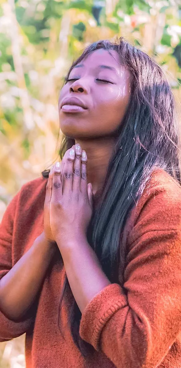 A woman with eyes closed and hands pressed together in a prayer or meditative pose outdoors, surrounded by blurred greenery.