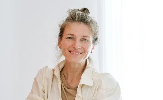 Smiling woman with gray hair tied back, wearing a beige top, in front of a white curtain