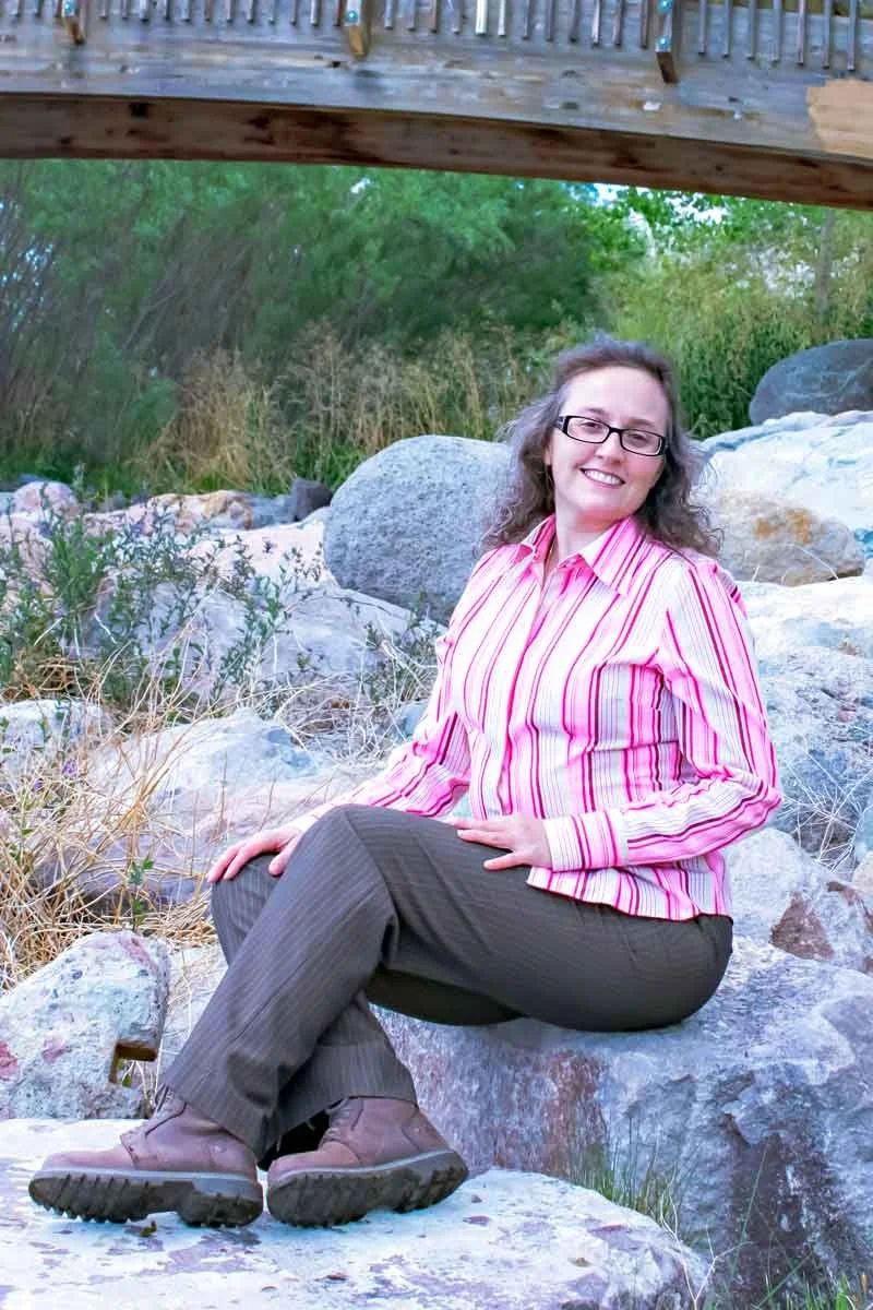 A woman sitting on rocks under a wooden bridge, smiling, wearing glasses, a pink striped shirt, and dark pants, surrounded by vegetation and rocks.