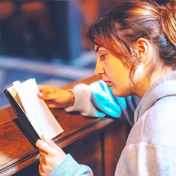 A young woman with brown hair tied back is sitting at a wooden table, reading a book.