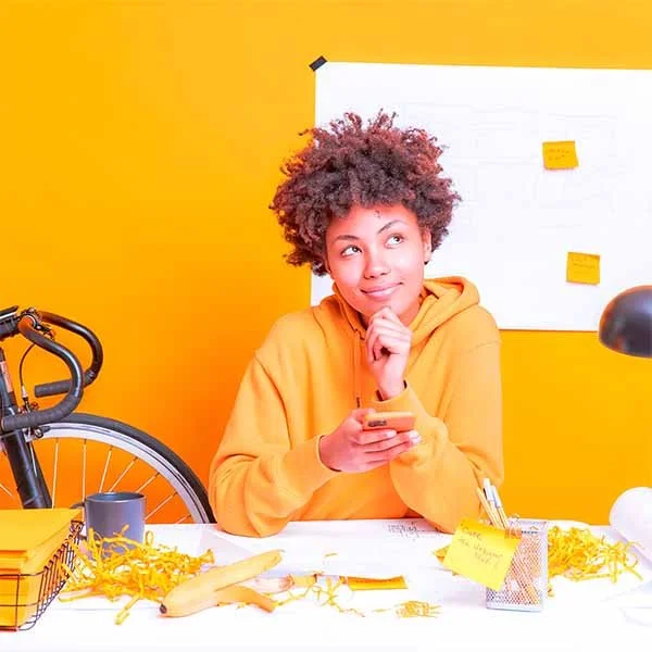 Young woman with curly hair in a yellow hoodie seated at a desk with yellow decor, holding a smartphone, surrounded by office supplies, items, and a bicycle in the background.