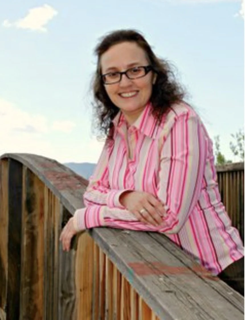 A woman with dark curly hair and glasses smiling, leaning on a wooden railing outdoors with a blue sky and clouds in the background.