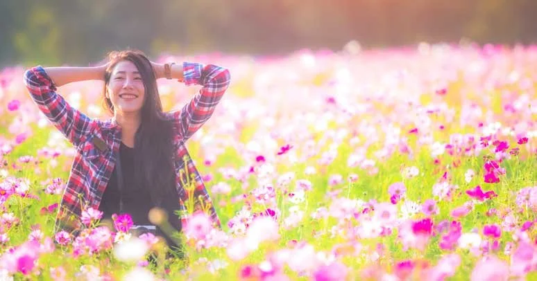 A woman smiling with arms behind her head sitting in a field of pink and white flowers